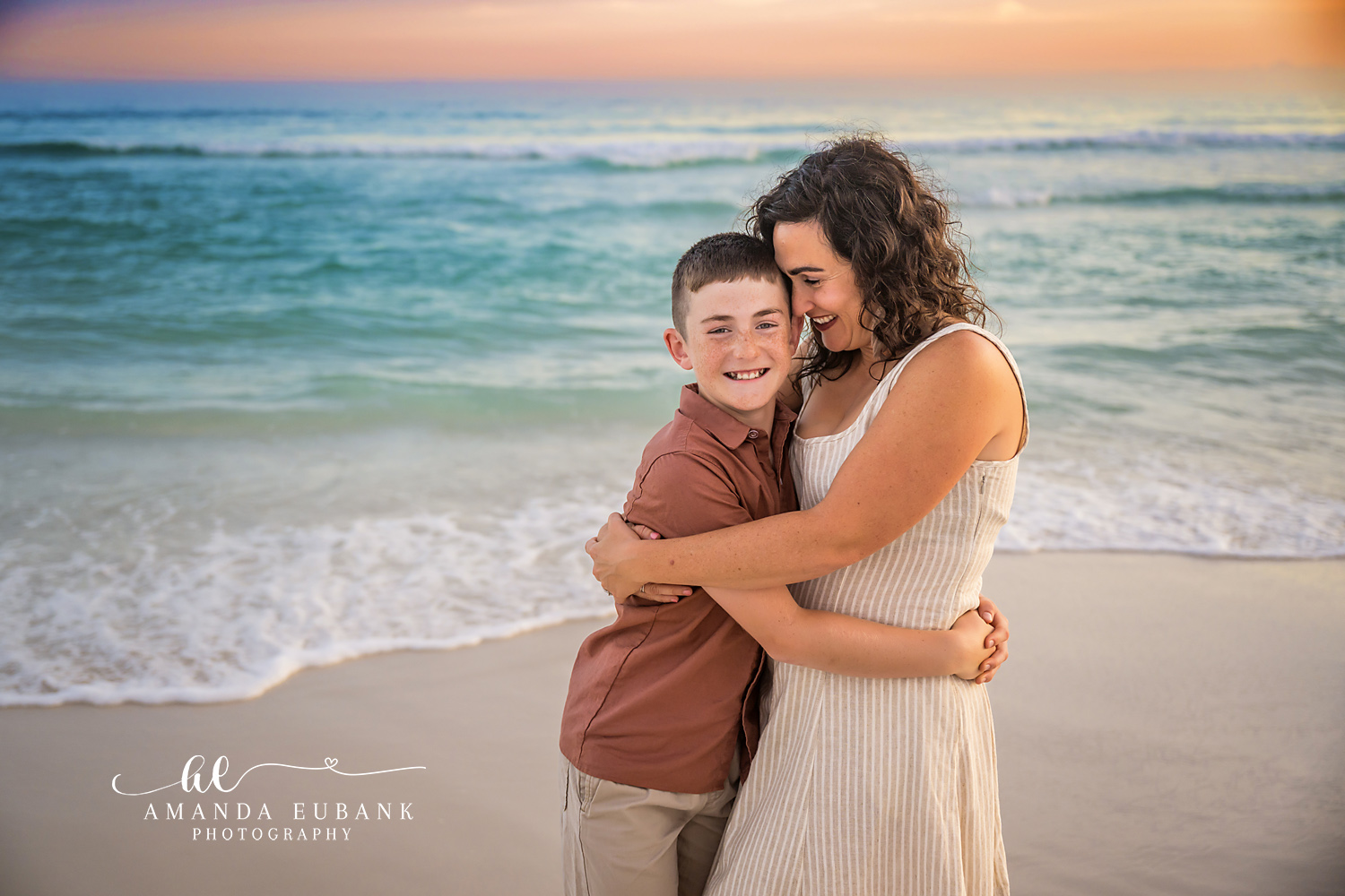 Destin Mother and Son Tender Sunset Portrait | Destin FL Family Photographer Mom kissing the top of her freckled sons head as he grins at the camera on the Destin FL beach at sunset with the Gulf and a soft pastel sky
