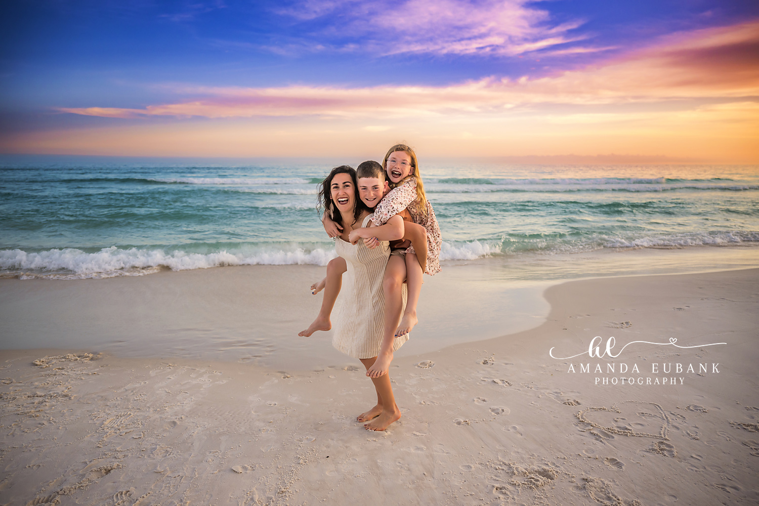 Destin Mom Piggybacking Both Kids Sunset Portrait | Destin FL Family Photographer Mom carrying both her son and daughter on her back all laughing on the Destin FL beach at sunset with the Gulf and a vivid orange and purple sky