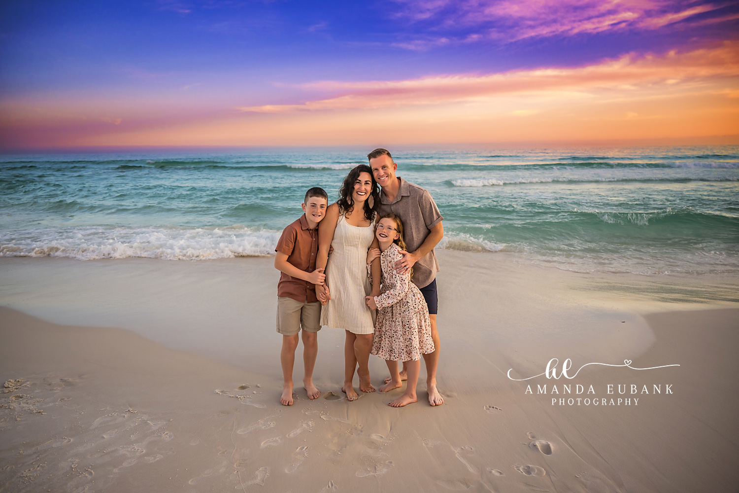 Destin Family of Four Sunset Shoreline Portrait | Destin FL Family Photographer Family of four gathered close on the Destin FL shoreline at sunset with the Gulf surf and a pastel orange purple and blue sky behind them