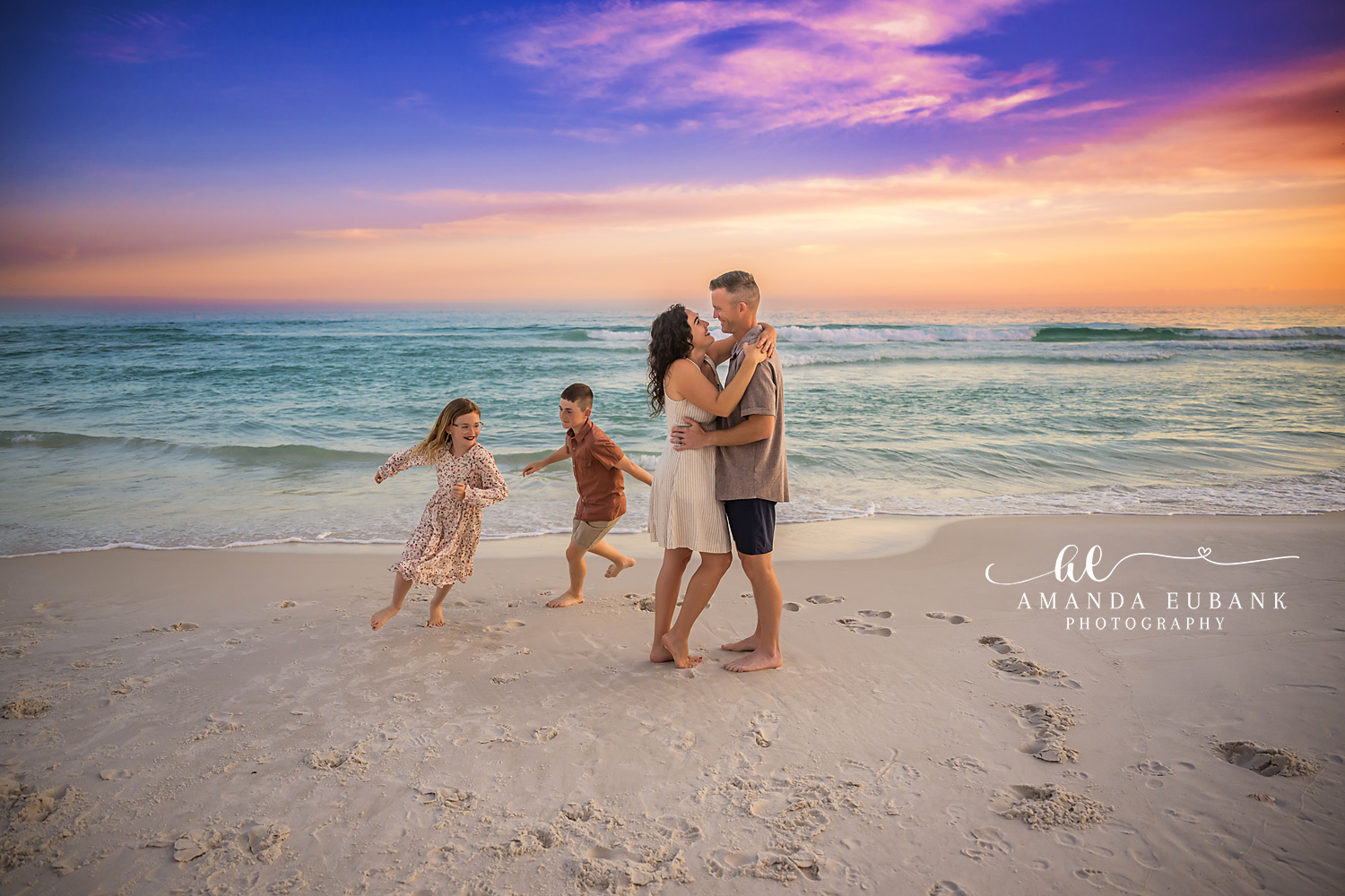Destin Parents Embrace Kids Running Sunset Portrait | Destin FL Family Photographer Mom and dad embracing while their son and daughter run around them on the Destin FL beach at sunset with the Gulf and a vivid purple and orange sky