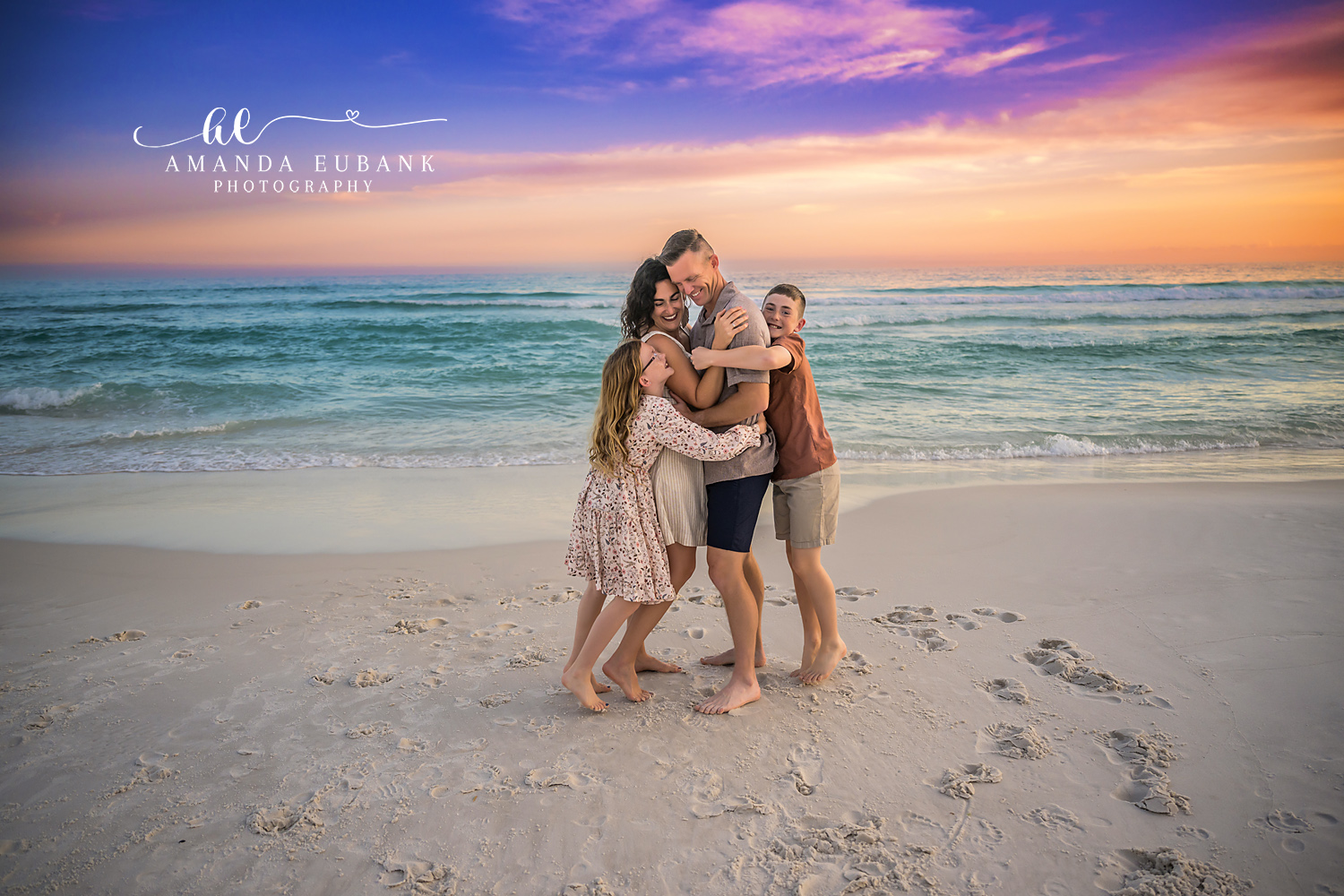 Destin Family Group Hug Sunset Portrait | Destin FL Family Photographer Family of four sharing a tight group hug on the Destin FL beach at sunset with the Gulf and a vivid purple and orange sky behind them