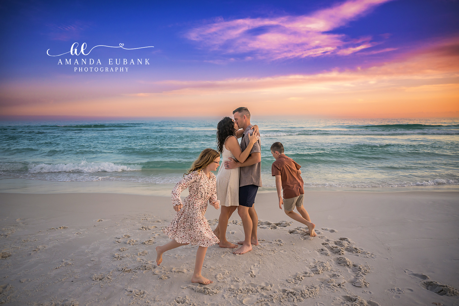 Mom and dad sharing a kiss while their daughter and son dance around them on the Destin FL beach at sunset with the Gulf and a pastel sunset sky