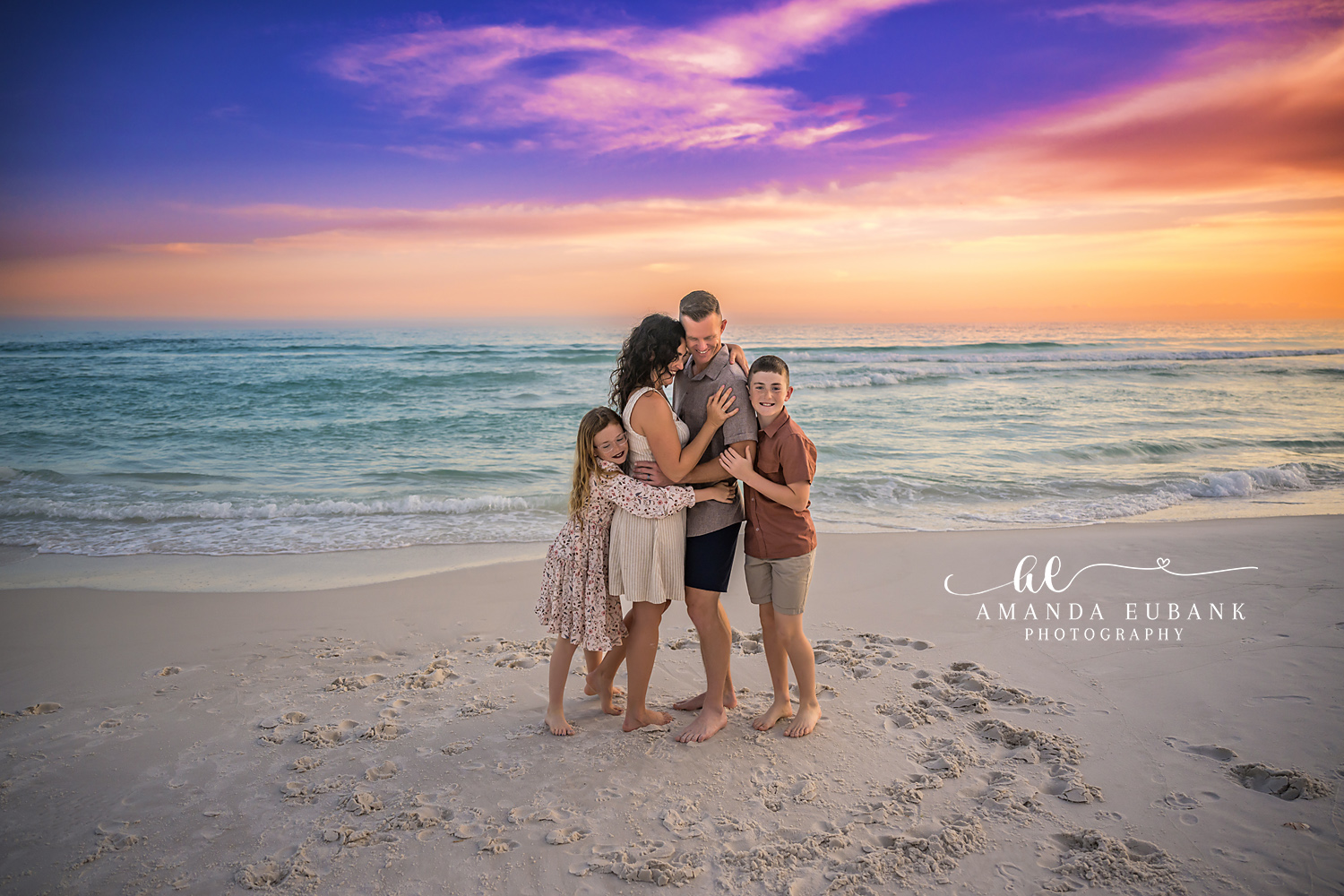 Destin Family Close Together Sunset Portrait | Destin FL Family Photographer Family of four standing together in a close warm embrace on the Destin FL beach at sunset with the Gulf and a vivid purple and orange sky behind them