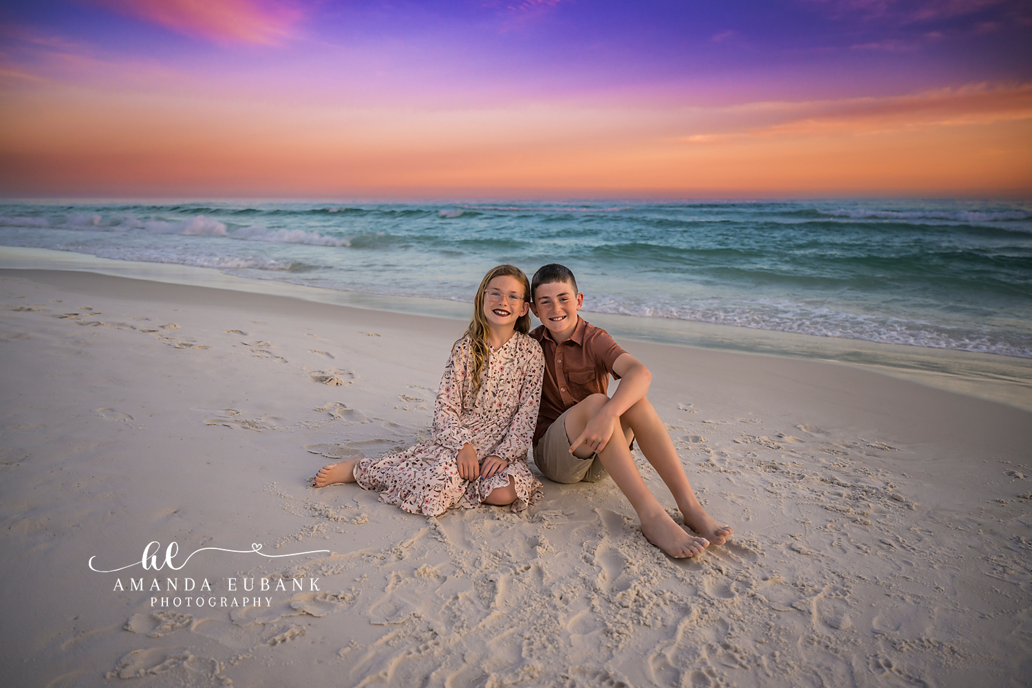 Destin Siblings Seated on the Sand Sunset Portrait | Destin FL Family Photographer Brother and sister seated on the white sand leaning into each other on the Destin FL beach at sunset with the Gulf and a pastel pink and orange sky