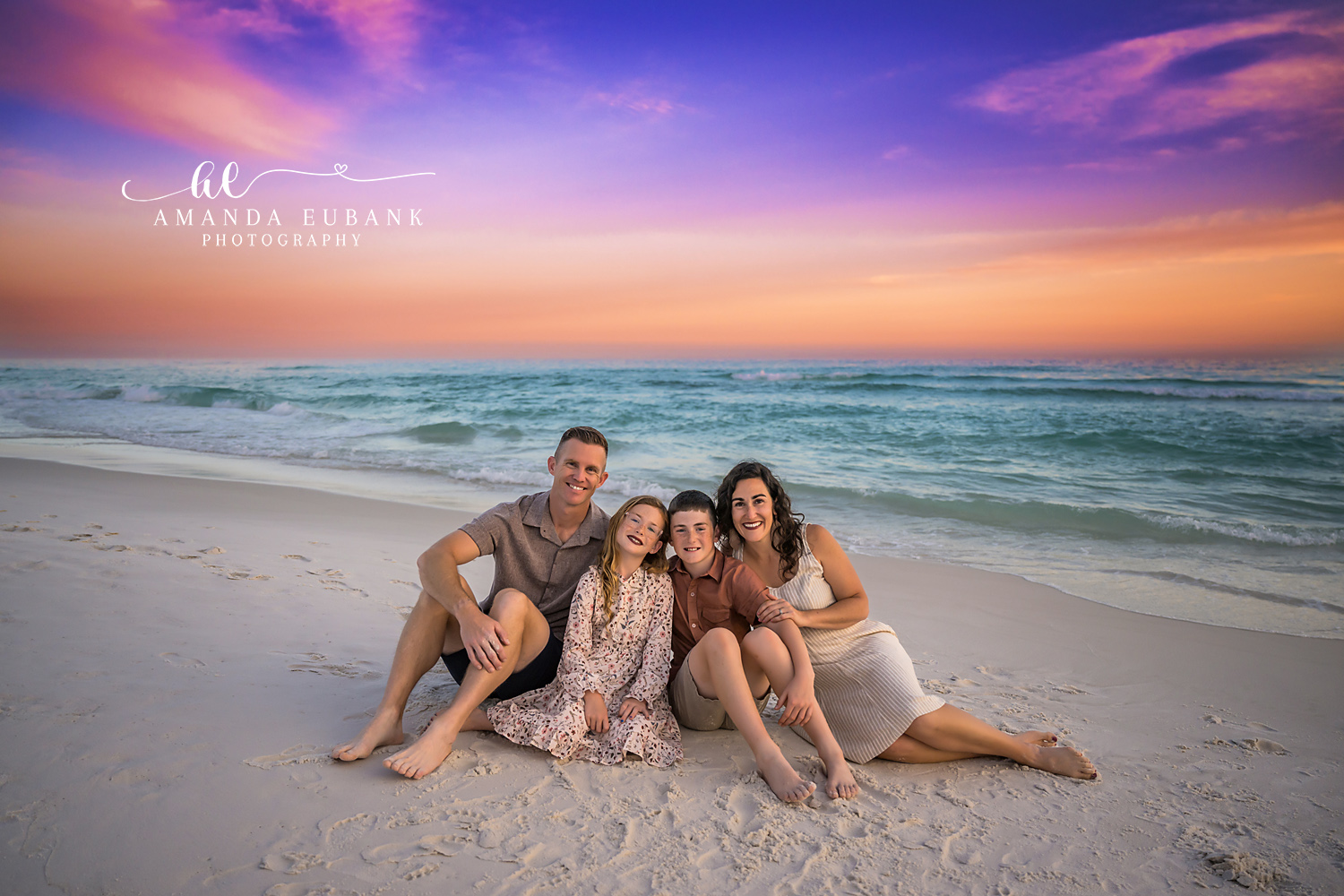 Destin Family Seated on the Sand Sunset Portrait | Destin FL Family Photographer Family of four seated together on the white sand of Destin FL at sunset with the Gulf and a pastel pink purple and orange sky behind them