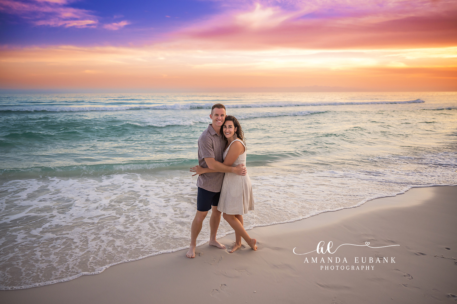 Destin Couple Sunset Shoreline Portrait | Destin FL Family Photographer Husband and wife embracing on the Destin FL shoreline at sunset with the Gulf surf and a vivid pink purple and orange sky behind them