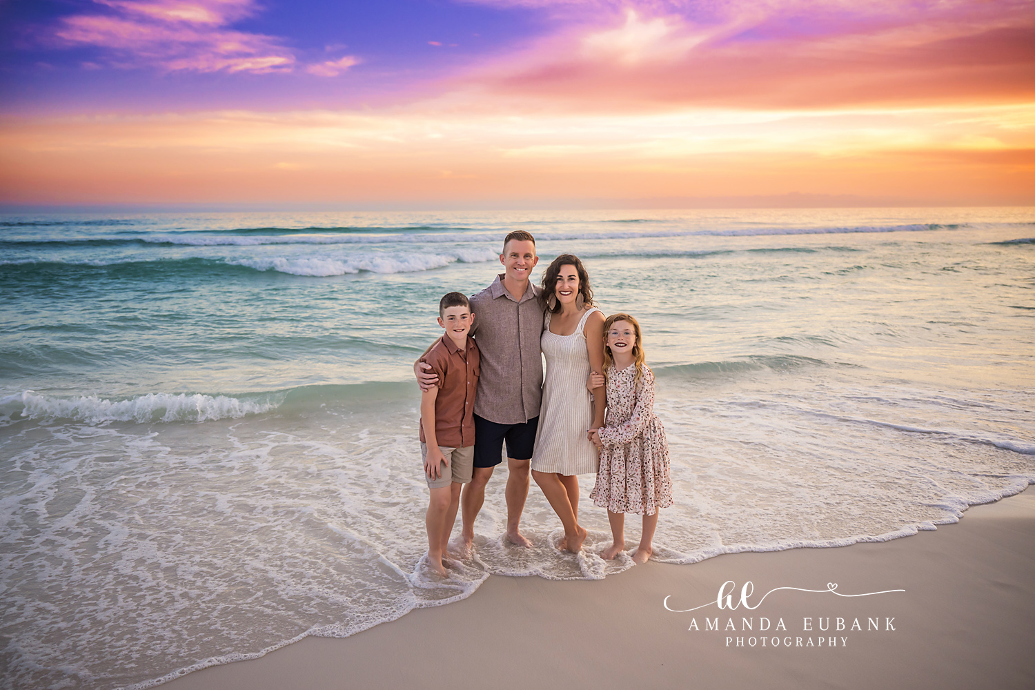 Family of four standing together in the foamy Gulf surf at Destin FL at sunset with a vivid purple and orange sky stretching behind them