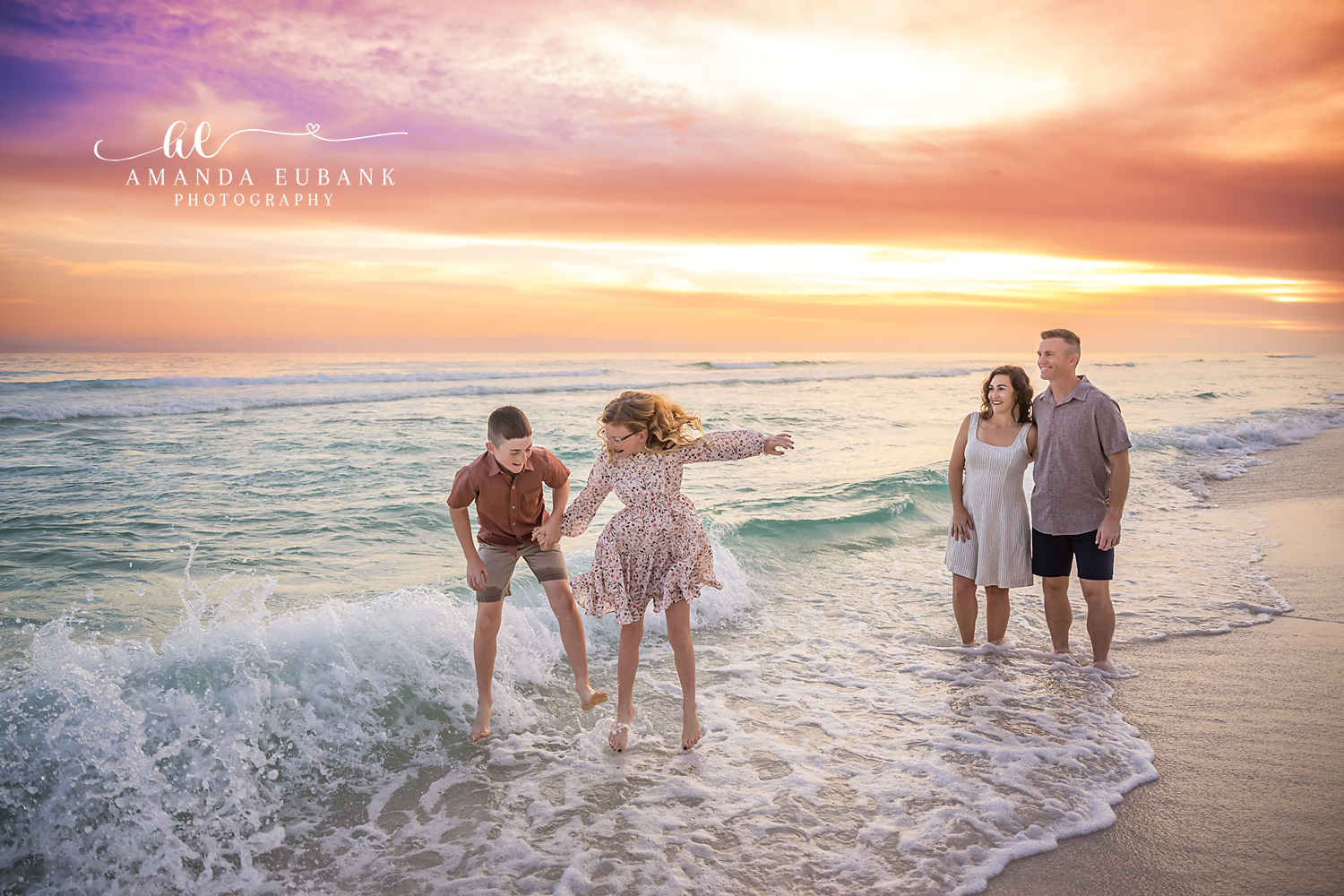 Destin Siblings Playing in the Surf with Parents Sunset | Destin FL Family Photographer Brother and sister playing in the foamy Gulf surf at Destin FL while their parents watch from behind beneath a vivid orange and pink sunset sky