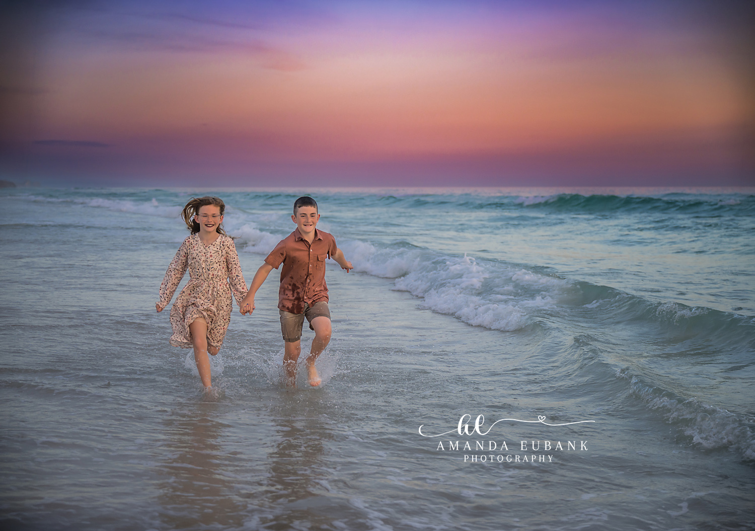 Destin Siblings Running Hand in Hand Through the Surf | Destin FL Family Photographer Brother and sister holding hands and running together through the foamy Gulf surf at Destin FL at sunset with a pastel pink and lavender sky