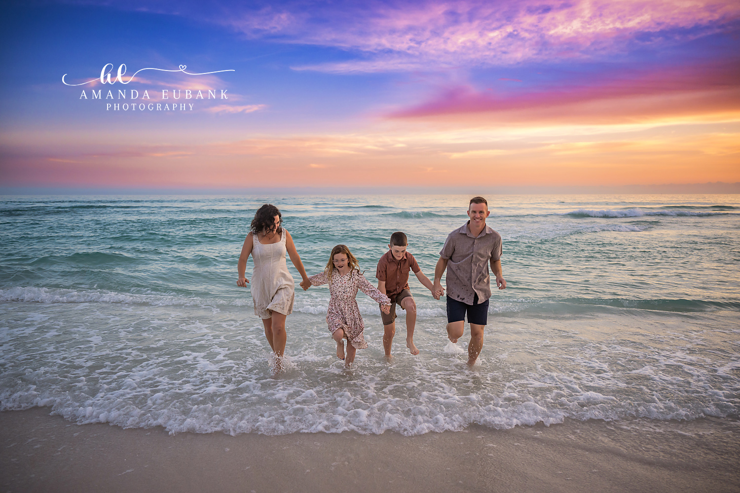 Destin Family Hand in Hand Through the Surf Sunset | Destin FL Family Photographer Family of four walking hand in hand through the foamy Gulf surf at Destin FL at sunset beneath a vivid purple pink and orange sky