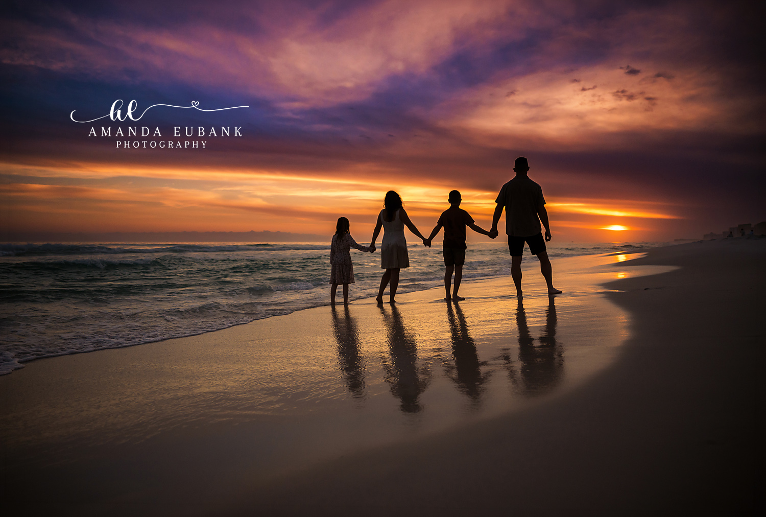 Silhouette of a family of four holding hands on the wet shoreline at Destin FL with the sun setting behind them and reflections in the sand