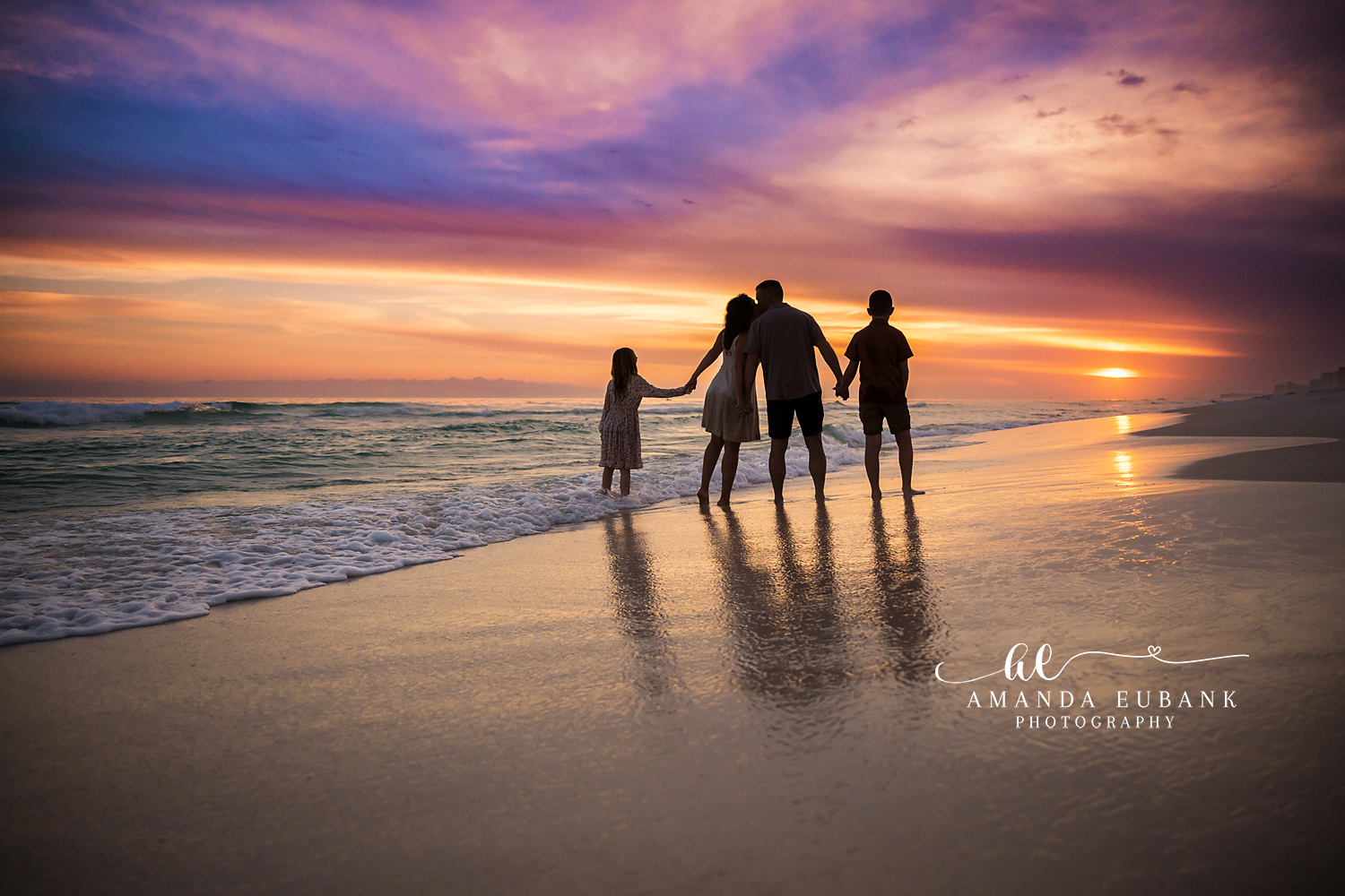 Destin Family Watching the Sunset Hand in Hand | Destin FL Family Photographer Family of four standing hand in hand watching the sun set over the Gulf at Destin FL with reflections in the wet shoreline beneath a fiery purple and orange sky