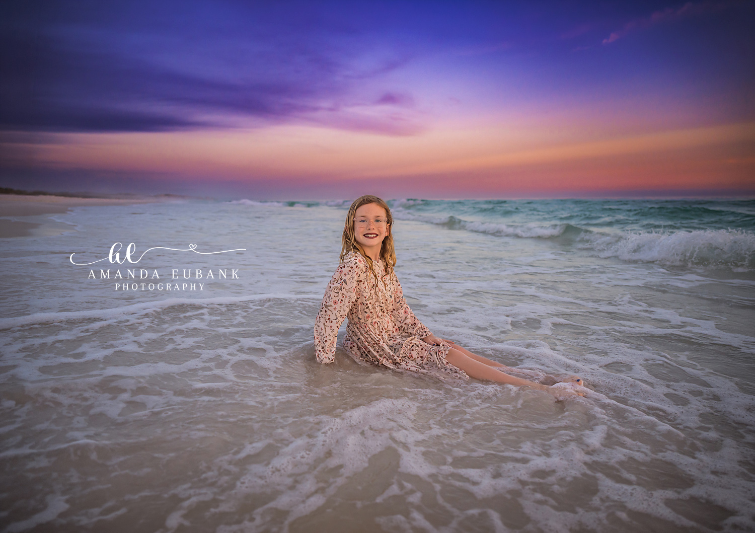 Destin Daughter Sitting in the Surf Sunset Portrait | Destin FL Family Photographer Red haired girl in a floral dress sitting in the foamy Gulf surf at Destin FL grinning at the camera beneath a pastel purple and pink sunset sky