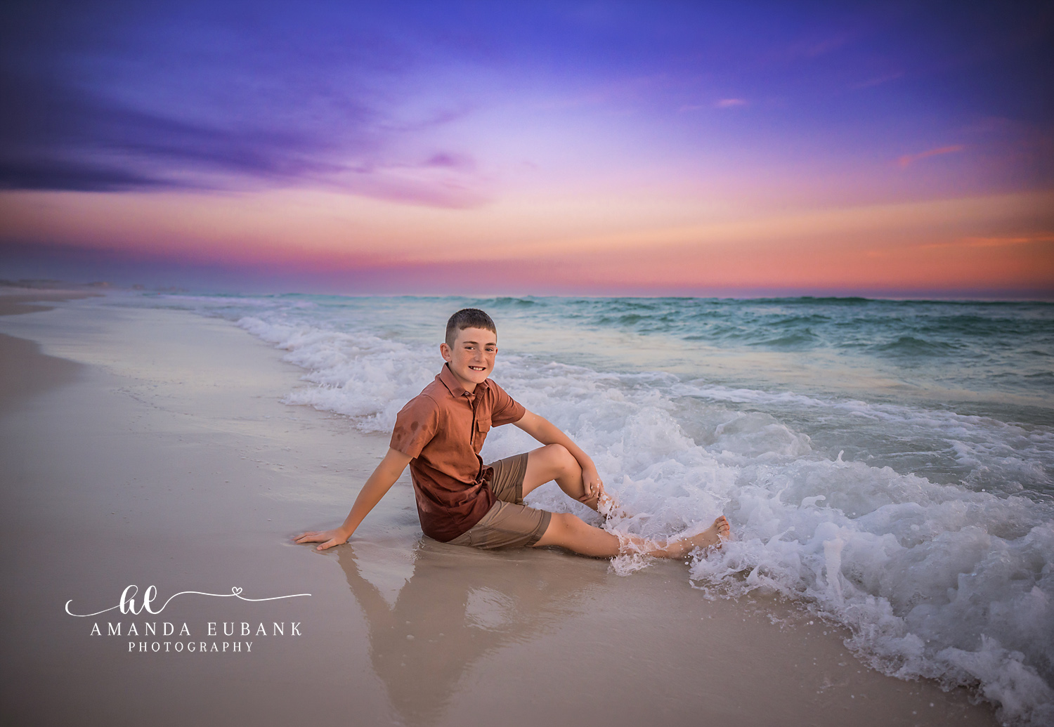Destin Son Sitting in the Surf Sunset Portrait | Destin FL Family Photographer Boy in a rust polo and cream shorts sitting in the foamy Gulf surf at Destin FL grinning at the camera beneath a pastel purple and pink sunset sky