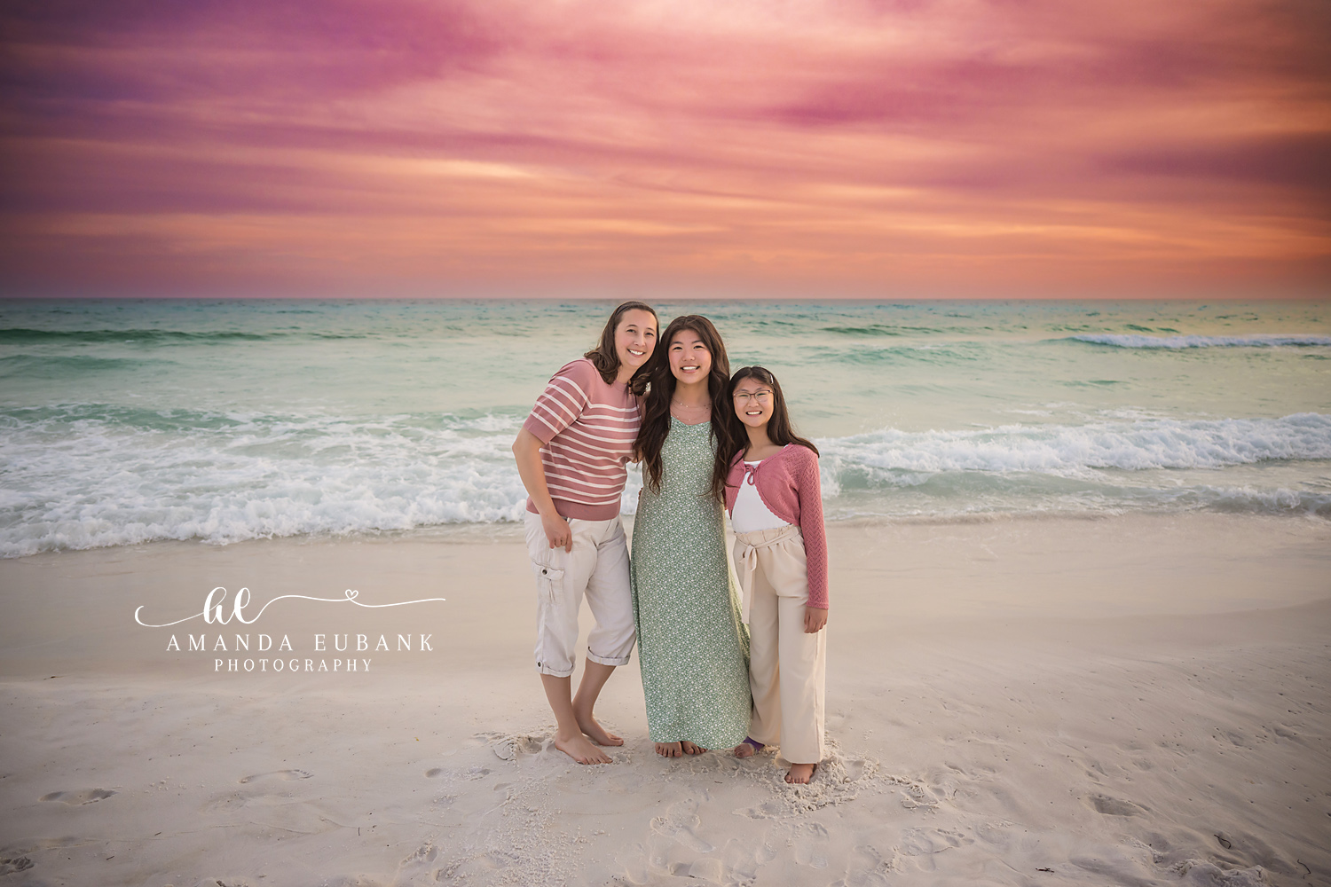 Miramar Beach Three Generations Sunset Portrait | Miramar Beach FL Family Photographer Three women of the M family in coordinated pink and sage green outfits standing barefoot on Miramar Beach FL sand at sunset with a vivid pink sky