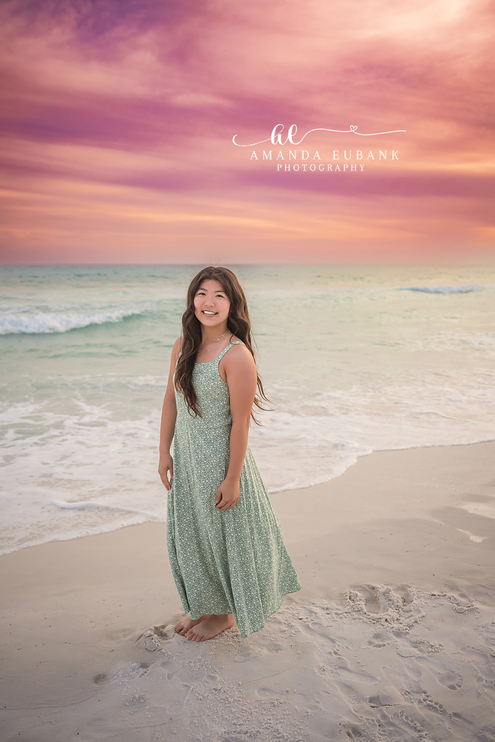 Miramar Beach Mom Sunset Portrait | Miramar Beach FL Family Photographer Mom in sage green floral maxi dress smiling over her shoulder barefoot on Miramar Beach FL sand at sunset with a pastel pink sky