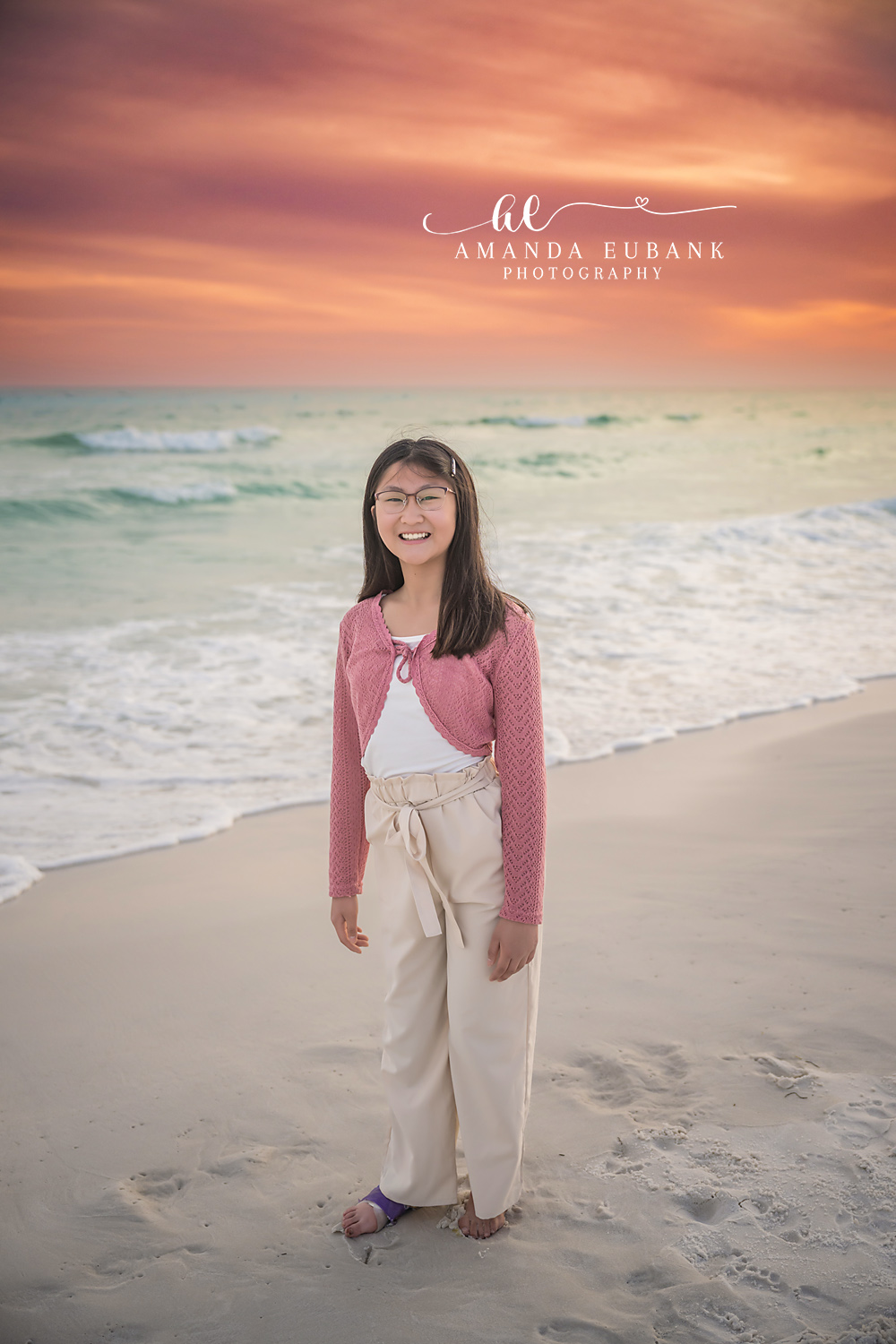 Miramar Beach Daughter Sunset Portrait | Miramar Beach FL Family Photographer Smiling girl in a pink crochet cardigan and cream wide-leg pants standing on Miramar Beach FL sand at sunset with the Gulf and a coral sky behind her