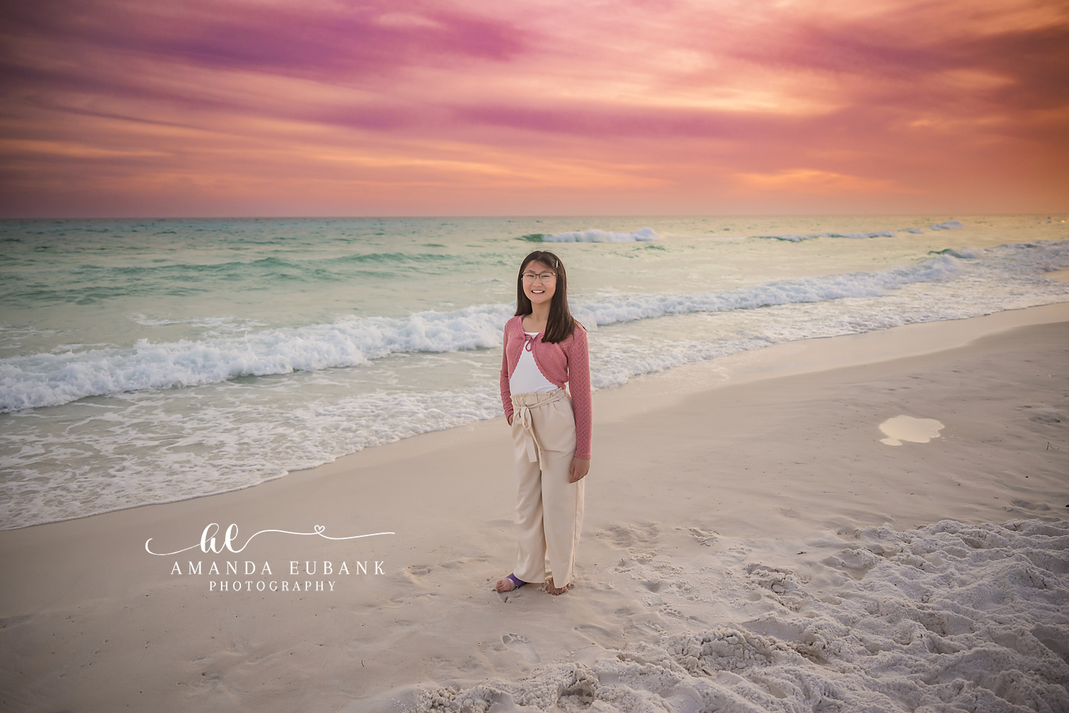 Miramar Beach Daughter Wide Sunset Sky Portrait | Miramar Beach FL Family Photographer Girl in pink crochet cardigan and cream pants standing on Miramar Beach FL with surf and a dramatic pink and orange sunset sky stretching overhead