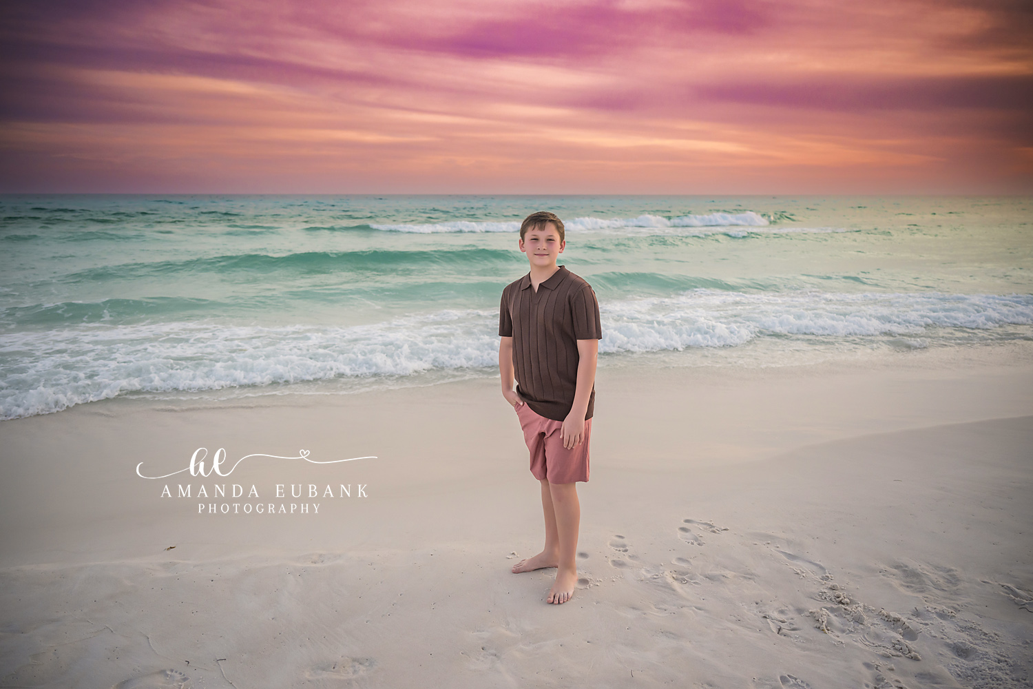 Miramar Beach Son Sunset Beach Portrait | Miramar Beach FL Family Photographer Young boy in a brown polo and coral shorts standing barefoot on Miramar Beach FL at sunset with the Gulf surf and a pink sky behind him