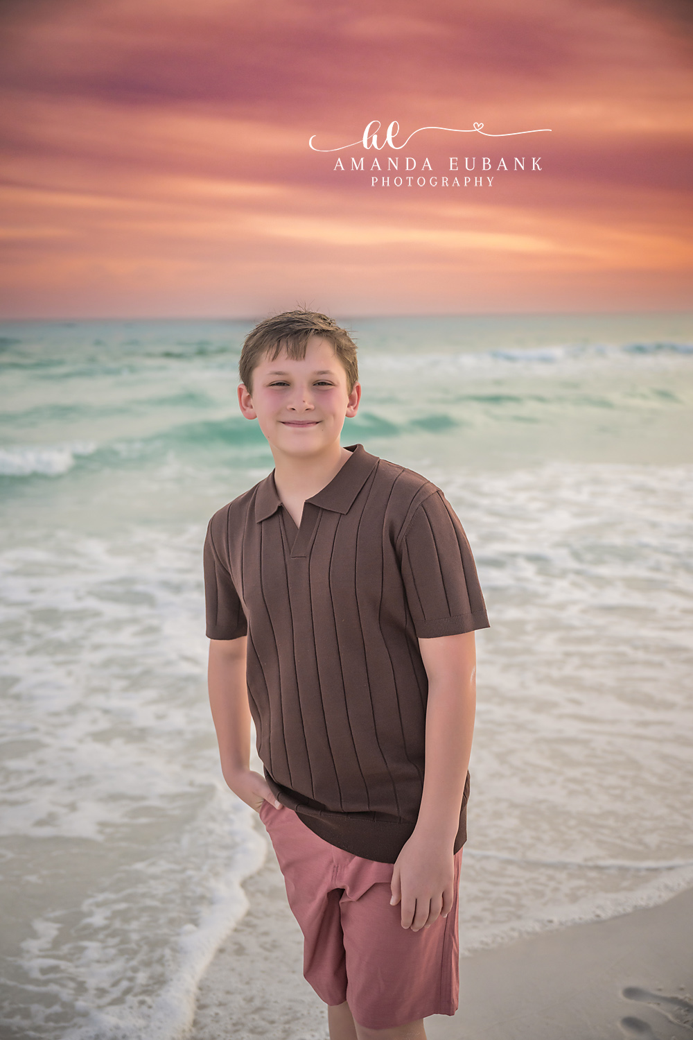Miramar Beach Son Casual Sunset Portrait | Miramar Beach FL Family Photographer Boy in brown polo and coral shorts smiling with one hand in his pocket on Miramar Beach FL at sunset with the Gulf surf behind him