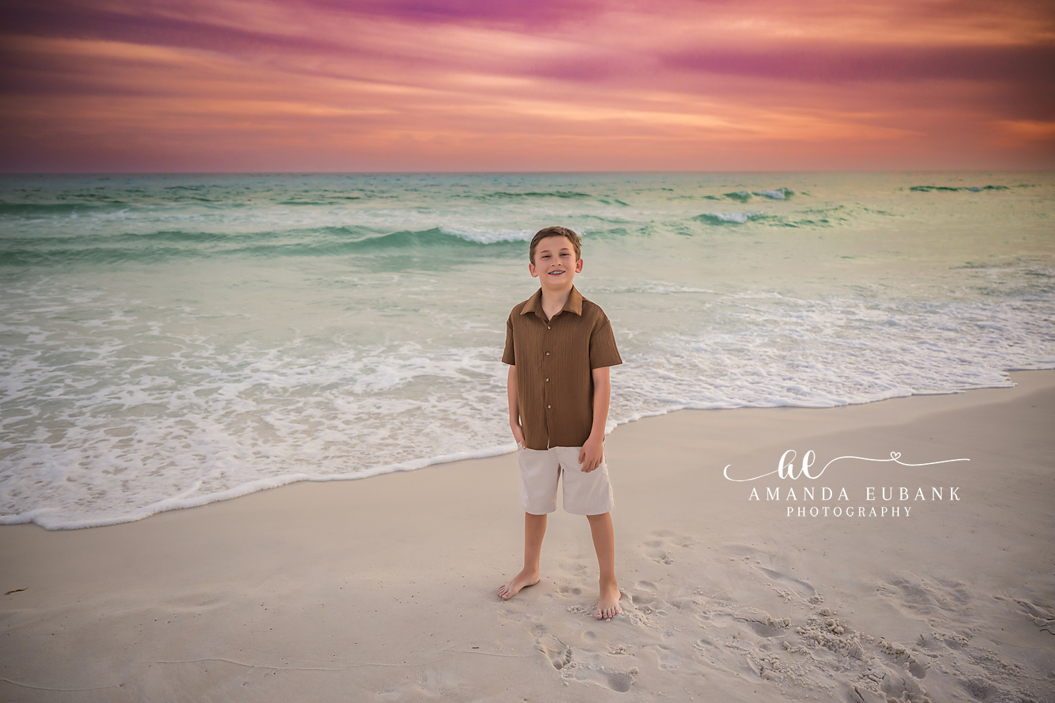Miramar Beach Youngest Son Sunset Beach Portrait | Miramar Beach FL Family Photographer Smiling young boy in a brown button-up shirt and cream shorts on Miramar Beach FL at sunset with the Gulf surf and a soft pink sky behind him