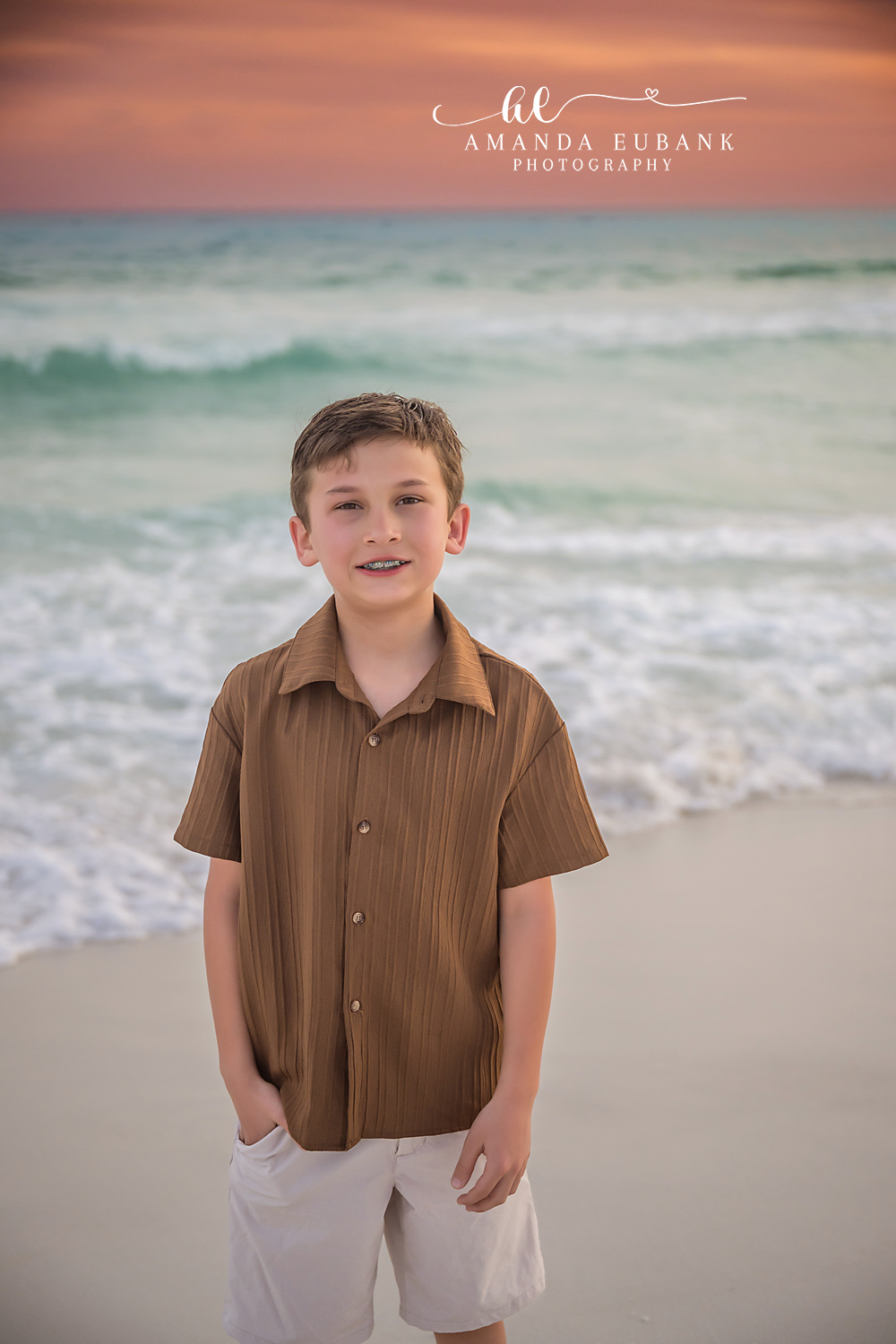 Miramar Beach Youngest Son Hands in Pockets Portrait | Miramar Beach FL Family Photographer Young boy in brown button-up shirt and cream shorts standing with hands in pockets on Miramar Beach FL at sunset with the Gulf surf behind