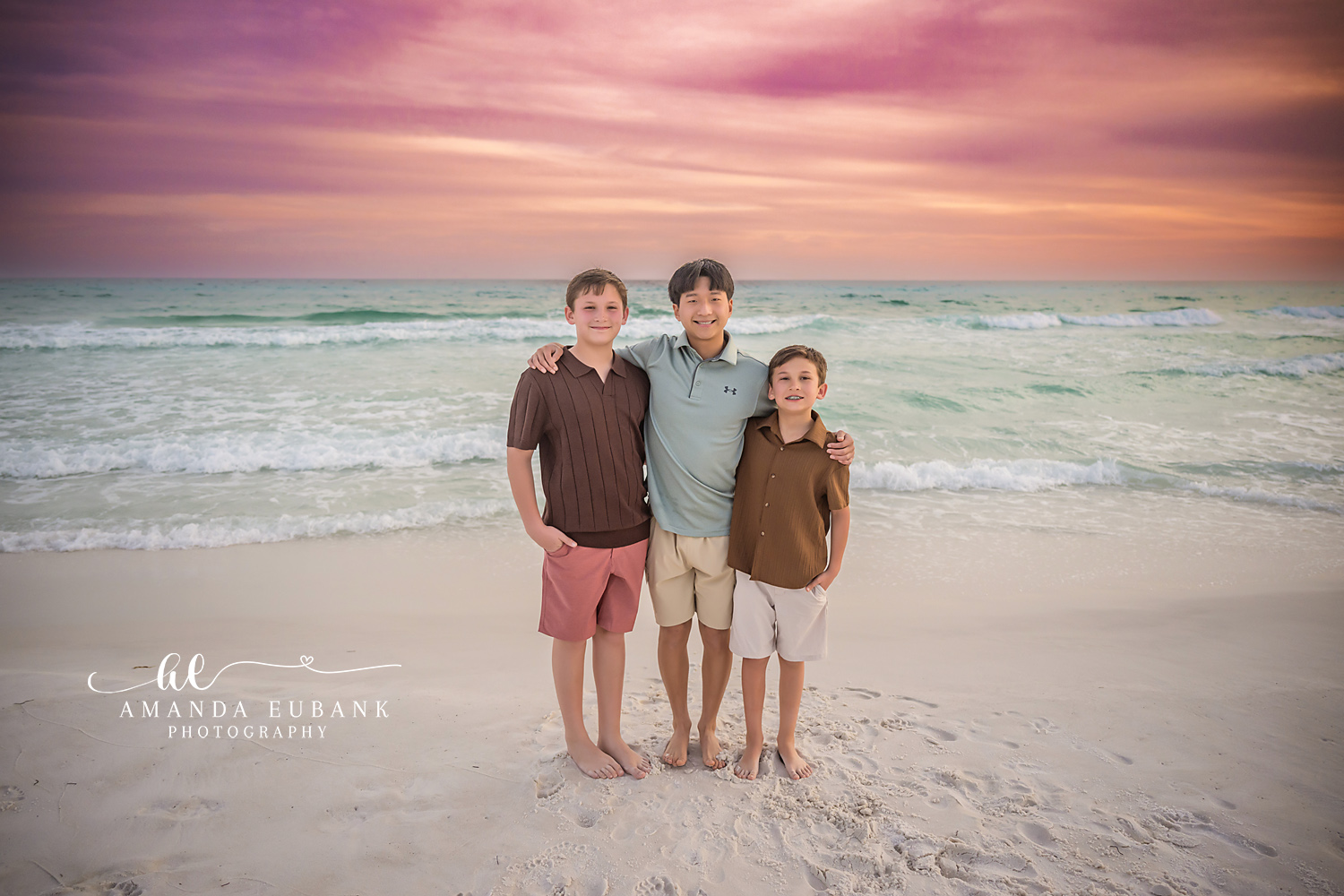 Miramar Beach Three Brothers Sunset Portrait | Miramar Beach FL Family Photographer Three brothers in coordinated brown sage and coral coastal outfits with arms around each other on Miramar Beach FL at sunset with surf and a pink sky