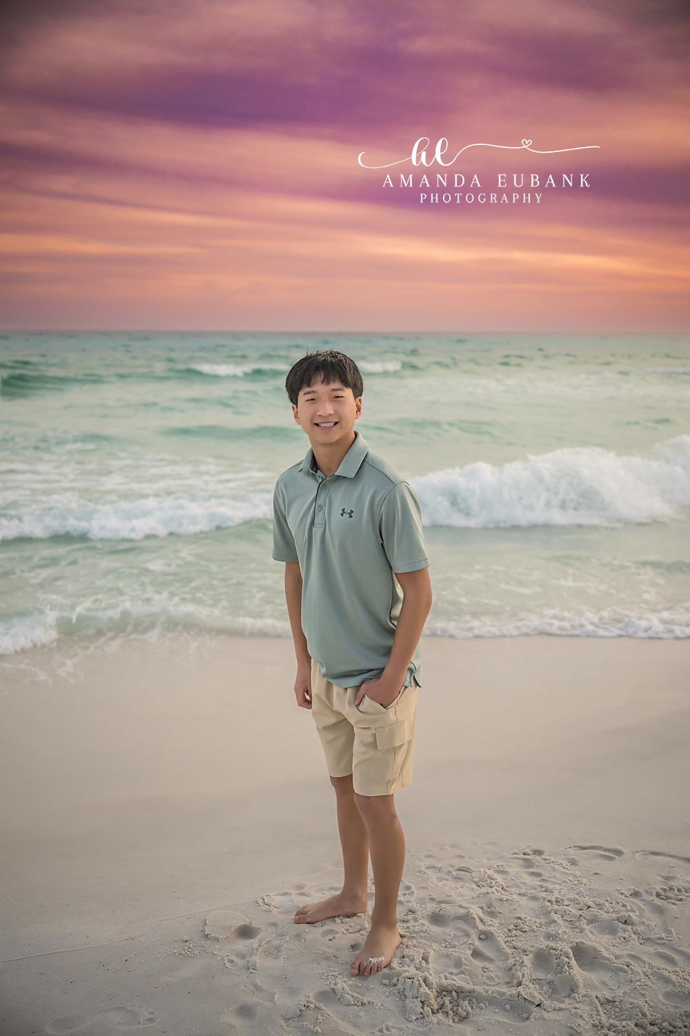 Miramar Beach Oldest Brother Sunset Portrait | Miramar Beach FL Family Photographer Older boy in a sage Under Armour polo and khaki shorts smiling on Miramar Beach FL at sunset with the Gulf surf and a purple pink sky behind him