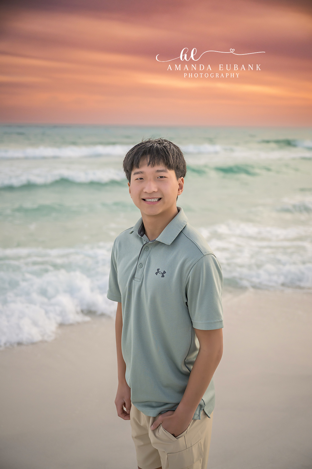 Miramar Beach Oldest Brother Close Up Sunset Portrait | Miramar Beach FL Family Photographer Close up sunset portrait of an older boy in a sage Under Armour polo smiling on Miramar Beach FL with the Gulf surf and a coral sky behind him