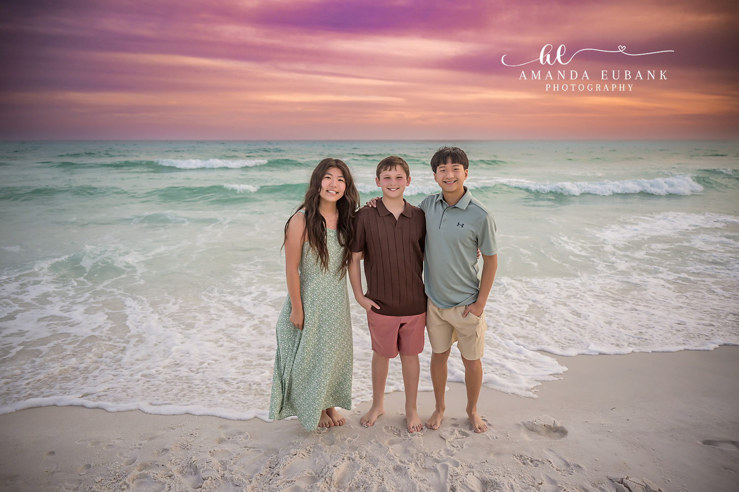 Miramar Beach Three Older Siblings Sunset Portrait | Miramar Beach FL Family Photographer Three older siblings in sage green pink coral and brown coastal outfits standing arm in arm on Miramar Beach FL at sunset with the Gulf surf and a pink sky