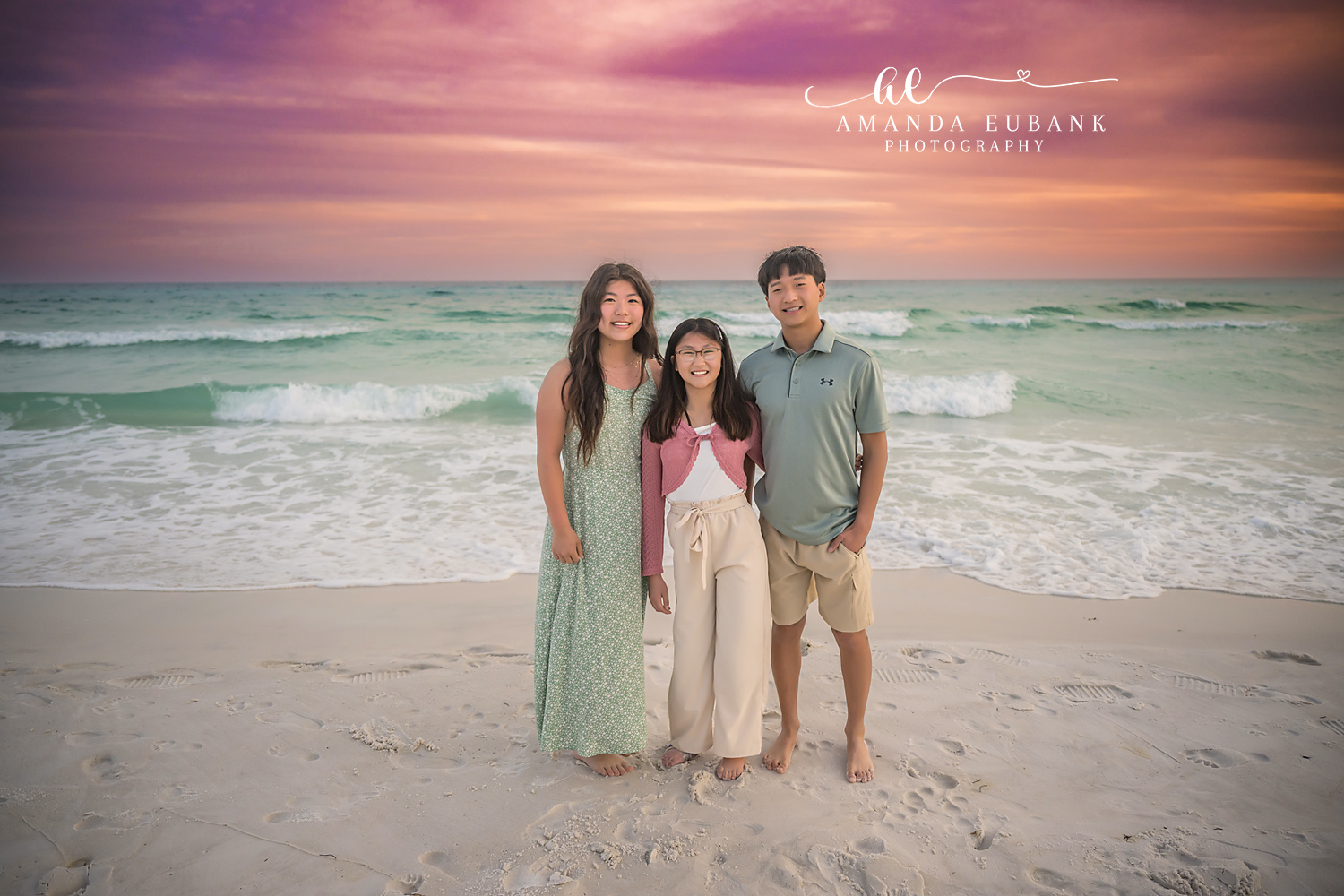Miramar Beach Sisters and Brother Sunset Portrait | Miramar Beach FL Family Photographer Two sisters and an older brother in coordinated sage pink and cream coastal outfits standing together on Miramar Beach FL at sunset with a soft pink sky