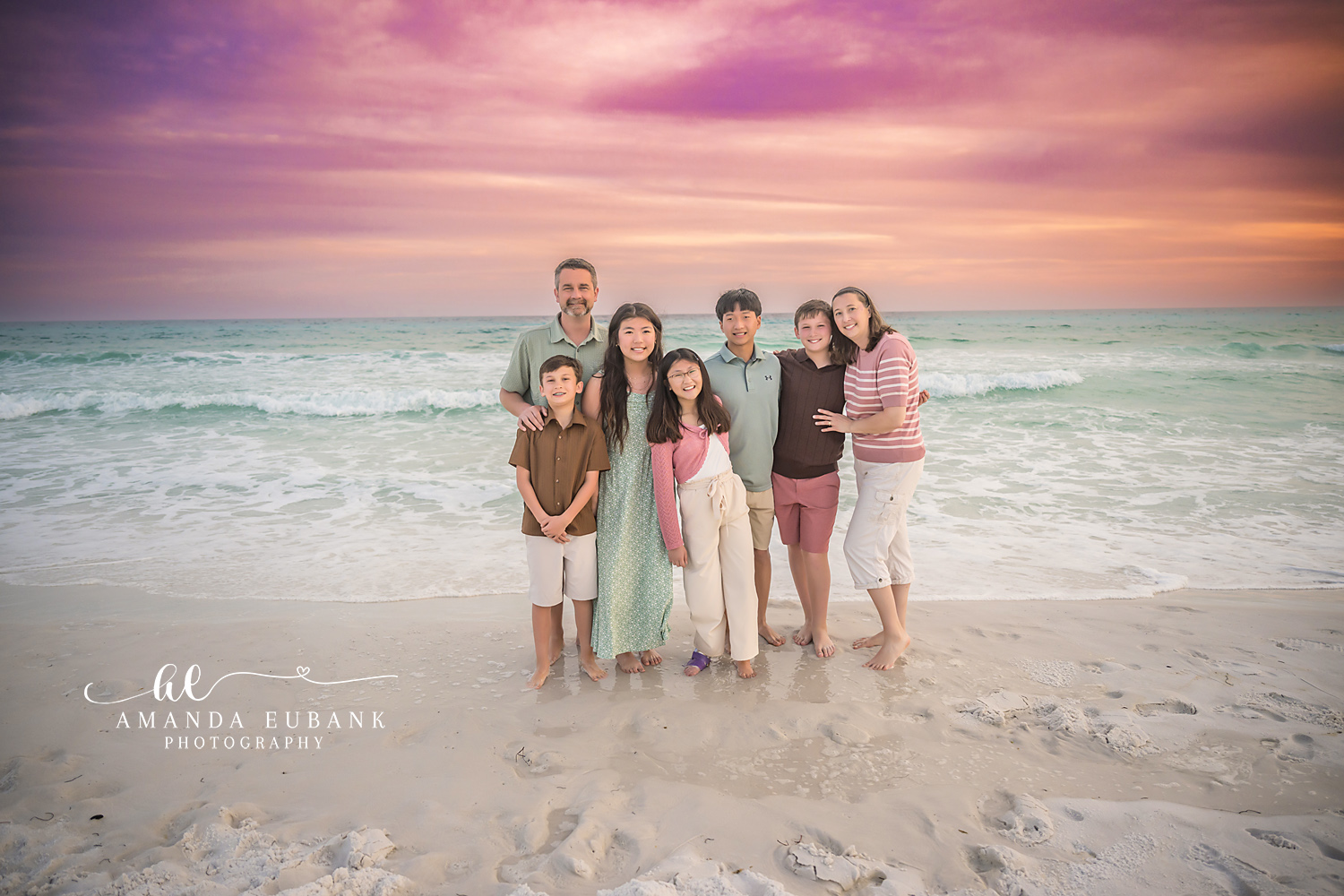Miramar Beach Family Huddle Sunset Portrait | Miramar Beach FL Family Photographer Family of seven gathered close in a warm huddle on Miramar Beach FL at sunset with a dramatic pink purple and orange sky behind them