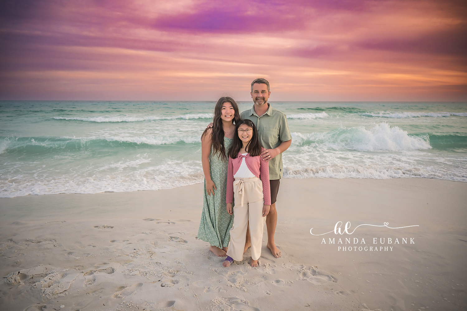 Miramar Beach Parents and Daughters Sunset Portrait | Miramar Beach FL Family Photographer Dad with his two daughters on Miramar Beach FL at sunset with the Gulf surf rolling in beneath a pink and purple sky