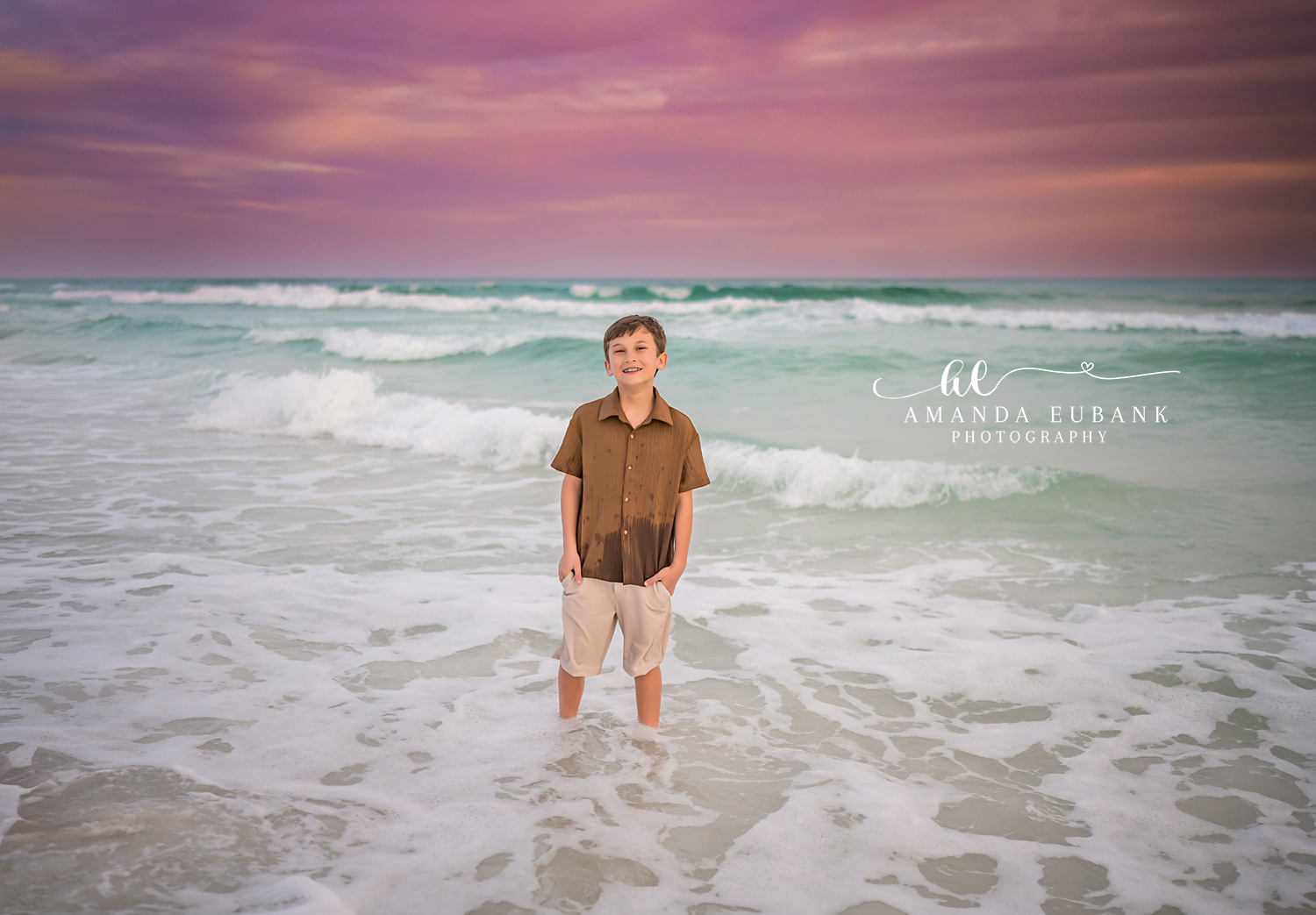 Miramar Beach Youngest Son in the Surf Sunset Portrait | Miramar Beach FL Family Photographer Young boy in a brown button up shirt and cream shorts standing in the foamy turquoise Gulf surf at Miramar Beach FL beneath a soft pink sky