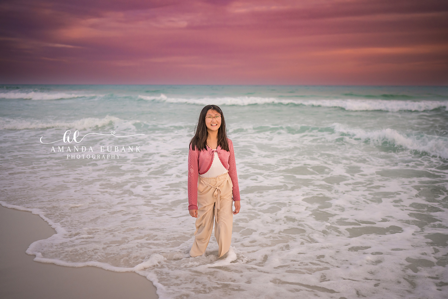 Miramar Beach Daughter in the Surf Sunset Portrait | Miramar Beach FL Family Photographer Smiling girl in a pink crochet cardigan and cream pants standing in the foamy Gulf surf at Miramar Beach FL beneath a soft pink and lavender sky