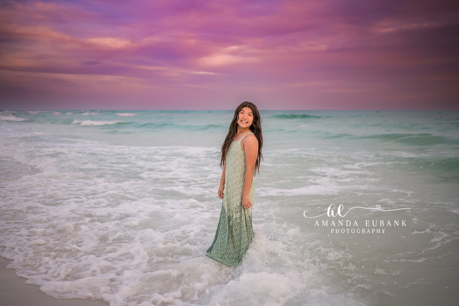 Miramar Beach Big Sister in the Surf Sunset Portrait | Miramar Beach FL Family Photographer Older sister in a sage green floral maxi dress standing in the foamy Gulf surf at Miramar Beach FL beneath a vivid purple and pink sunset sky