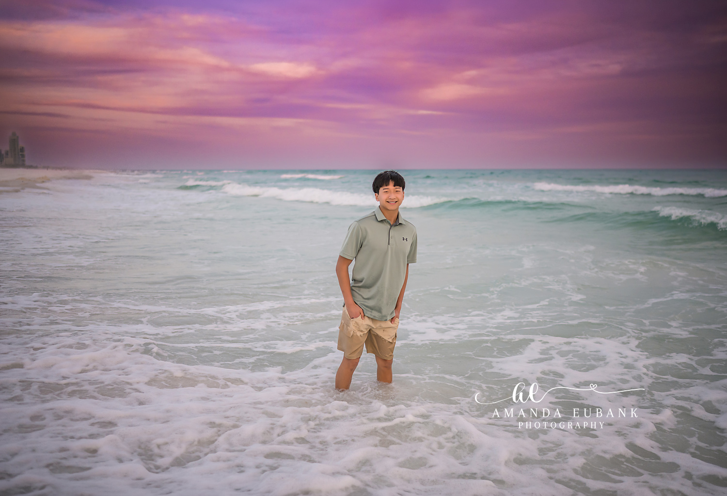 Miramar Beach Big Brother in the Surf Sunset Portrait | Miramar Beach FL Family Photographer Older boy in a sage Under Armour polo and khaki shorts standing in the foamy Gulf surf at Miramar Beach FL beneath a vivid pink and purple sky
