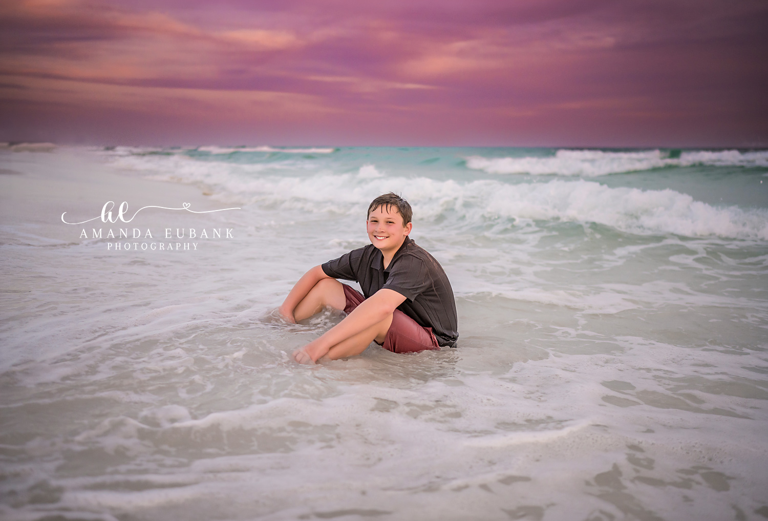 Miramar Beach Son Sitting in the Surf Sunset Portrait | Miramar Beach FL Family Photographer Boy in a brown polo and coral shorts grinning while sitting in the foamy Gulf surf at Miramar Beach FL with a soft pink and lavender sunset sky