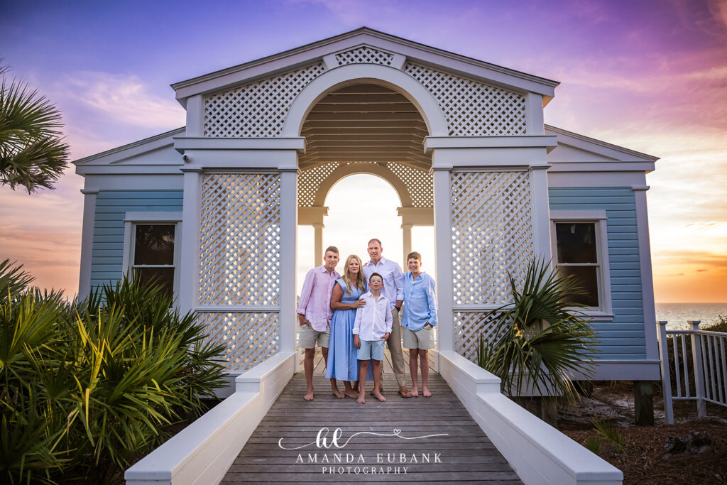 Family of five posed under the white Seaside, Florida boardwalk pavilion as a pastel pink and purple sunset glows over the Gulf of Mexico.