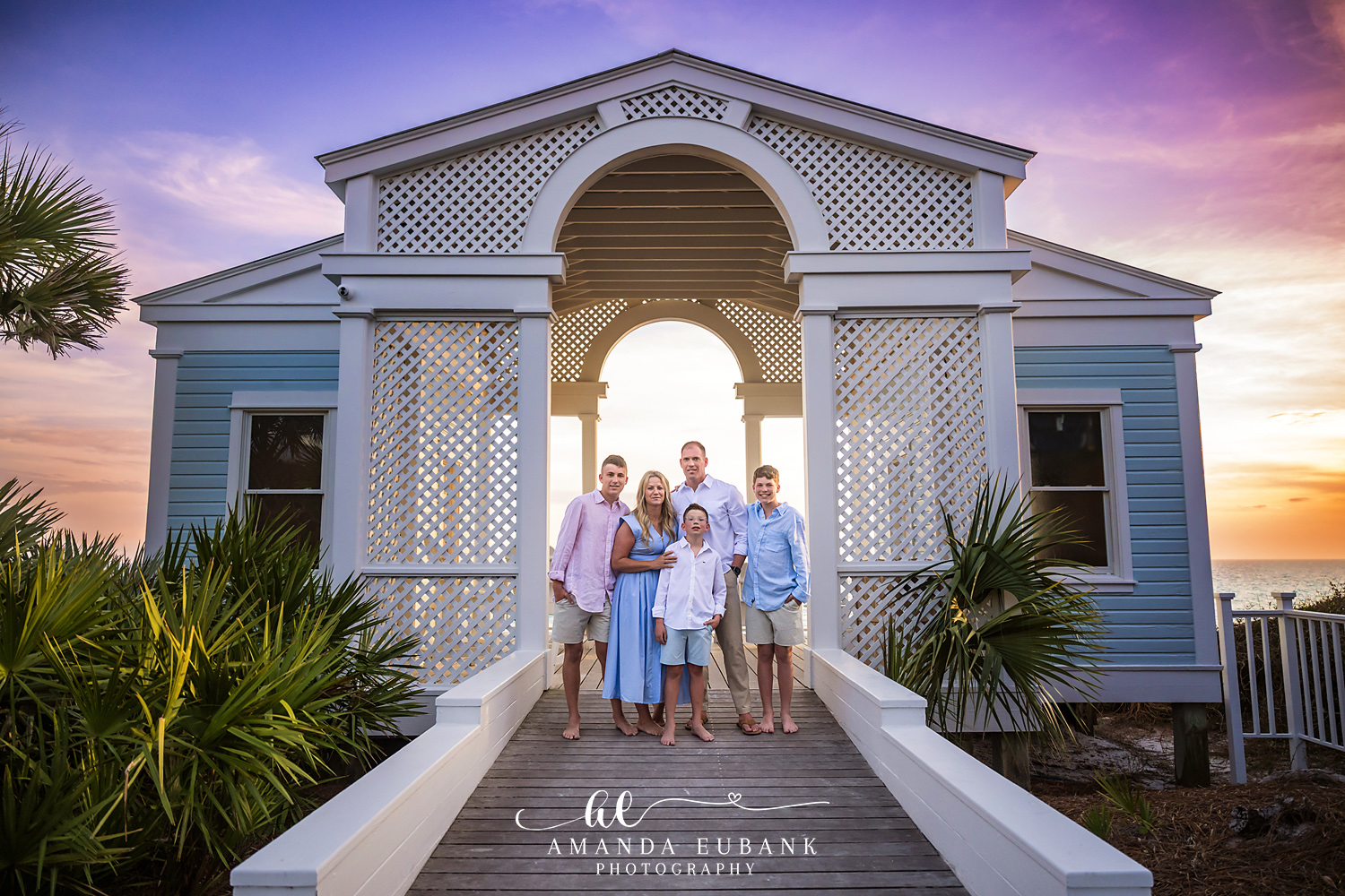 Seaside FL Family Photographer | Pavilion Boardwalk Sunset Portrait Family of five posed under the white Seaside, Florida boardwalk pavilion as a pastel pink and purple sunset glows over the Gulf of Mexico.