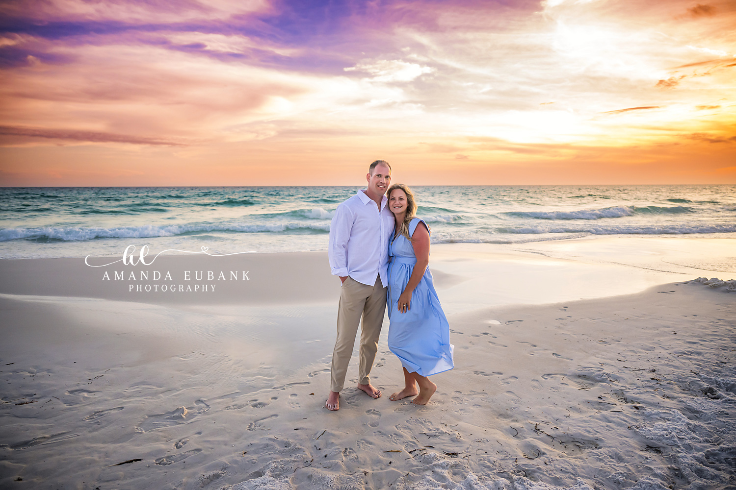 Seaside FL Couples Portrait | Sunset on the Gulf Shoreline Husband and wife in white and cornflower blue cuddle barefoot on the Seaside, Florida sand as a dramatic orange and pink sunset glows behind the Gulf surf.