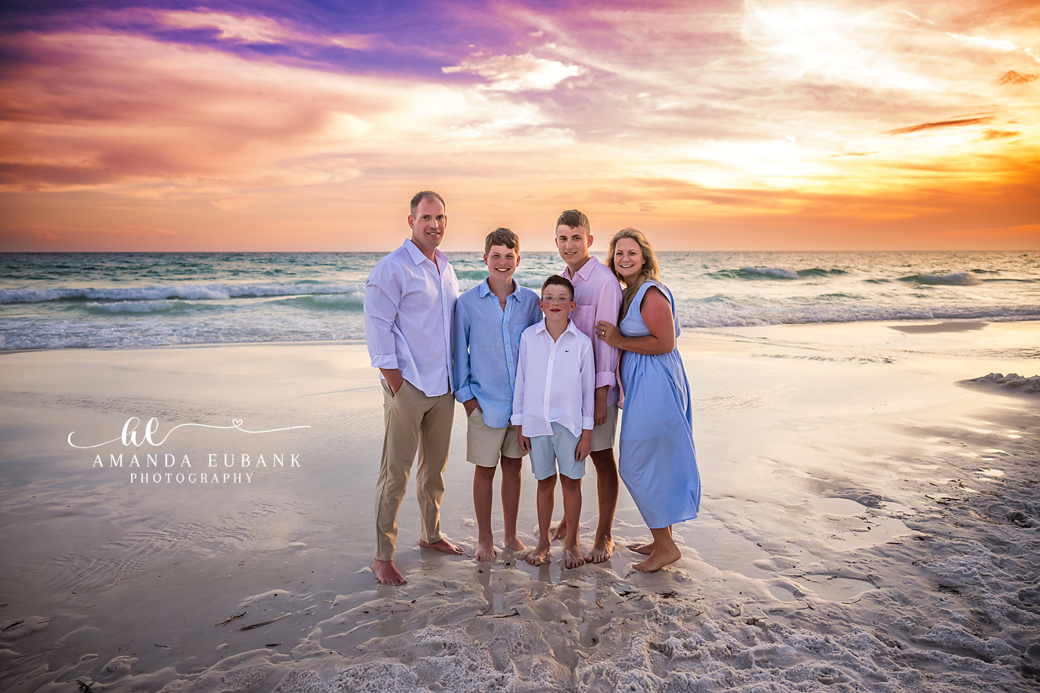 Seaside FL Family of Five Sunset Portrait | Gulf Shoreline Family of five in coordinated white, pink, and cornflower blue stand together on the wet sand at the Seaside, FL shoreline as a brilliant orange and lavender sunset glows over the Gulf.