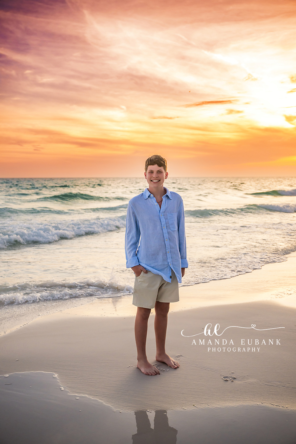 Seaside FL Tween Boy Sunset Portrait | Gulf Shoreline Solo Smiling tween boy in a light blue button-up and khaki shorts stands barefoot on the wet Seaside, FL sand at sunset, brilliant orange sky and Gulf surf behind him.