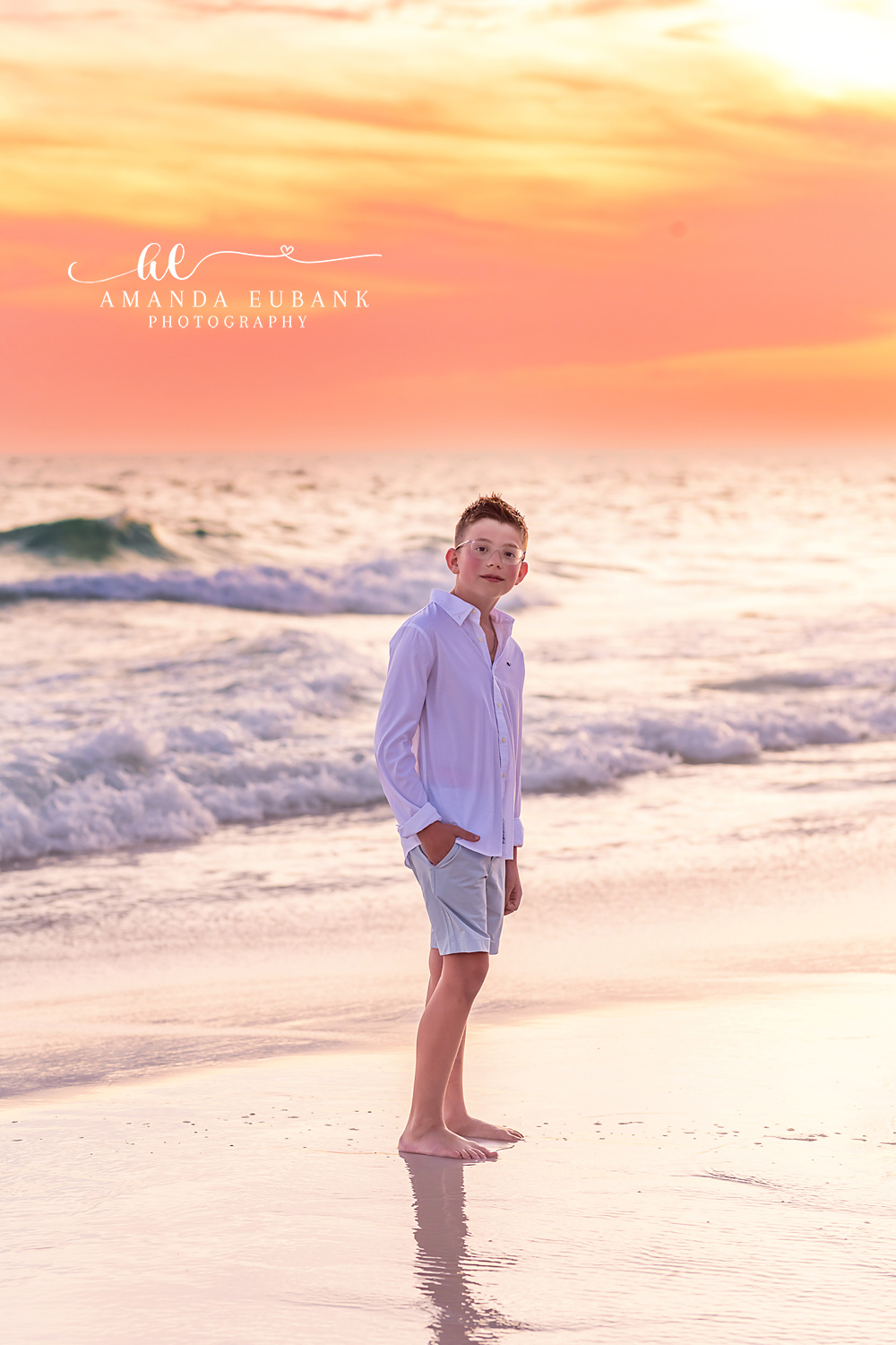 Seaside FL Young Boy Sunset Portrait | Solo on the Gulf Shoreline Young boy in a soft pink button-up and grey-blue shorts glances back over his shoulder while standing barefoot at the Seaside, FL surf line beneath a peach pastel sunset.