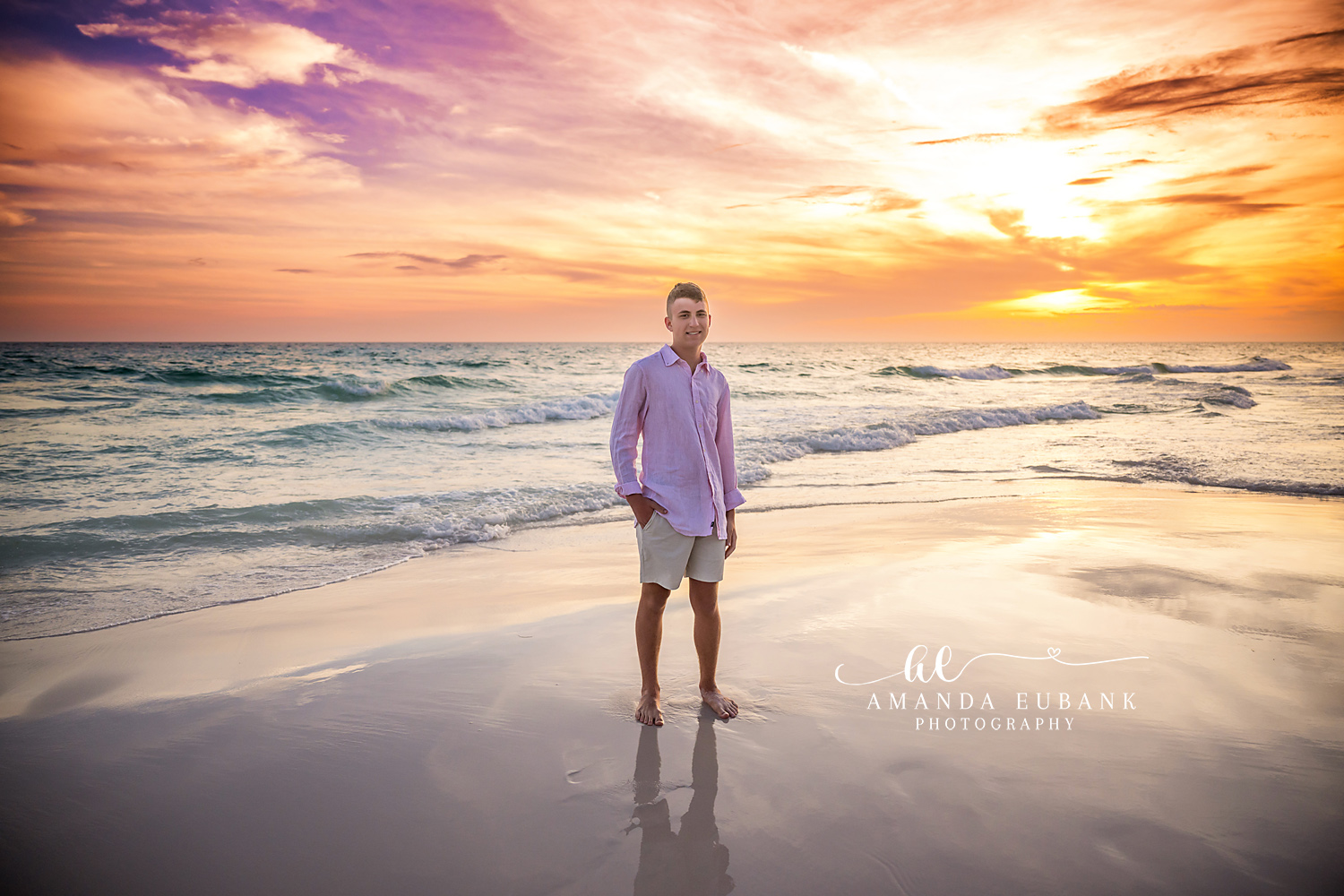Seaside FL Senior Portrait | Oldest Brother at Gulf Sunset Senior-aged young man in a soft pink button-up and khaki shorts stands barefoot on the wet Seaside, FL sand as a dramatic purple, orange, and gold sunset paints the Gulf horizon.