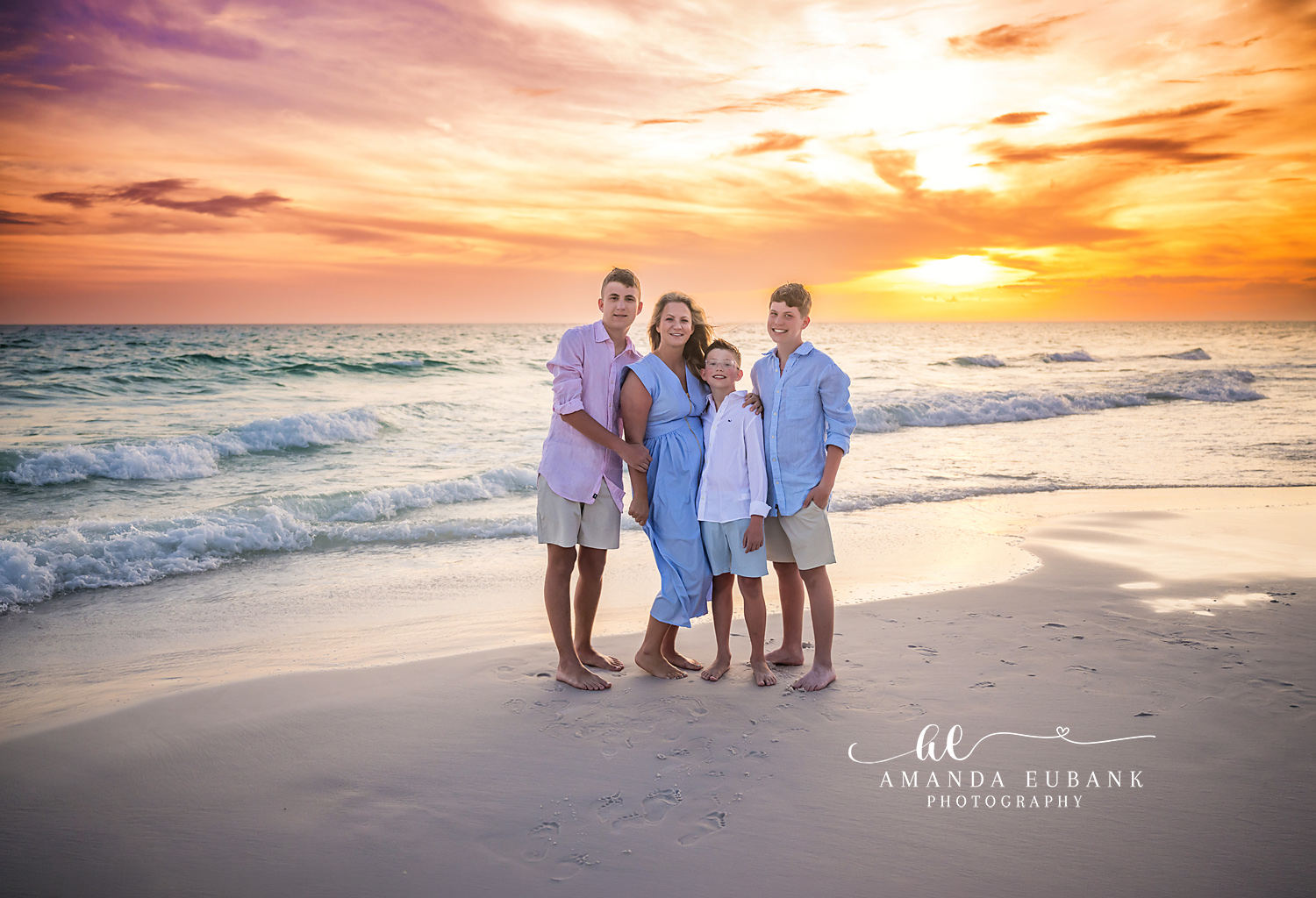 Seaside FL Mother and Sons Sunset Portrait | Boys Mom Beach Session Mother in a cornflower blue dress hugged by her three sons in pink, white, and light blue at the foamy Seaside, FL surf line beneath a glowing orange and lavender sunset.
