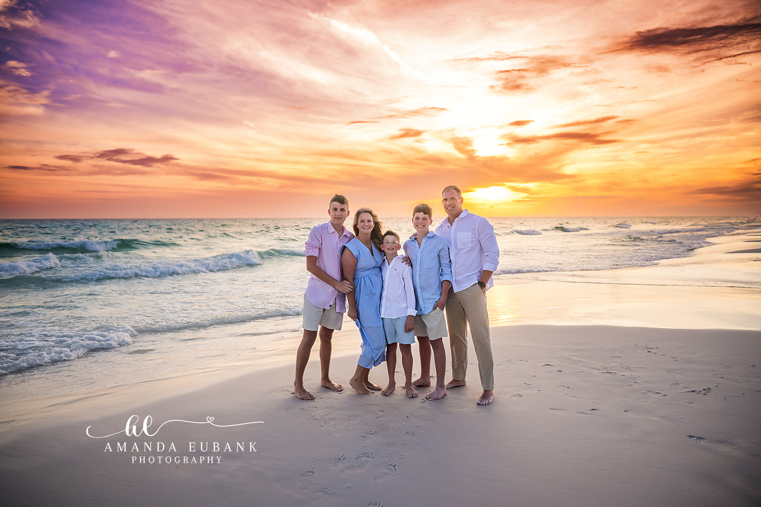 Seaside FL Family of Five Sunset Beach Portrait | 30A Photographer Family of five gathered along the foamy Seaside, FL surf line in coordinated pink, blue, and white as a vivid purple, orange, and gold sunset bursts over the Gulf horizon.