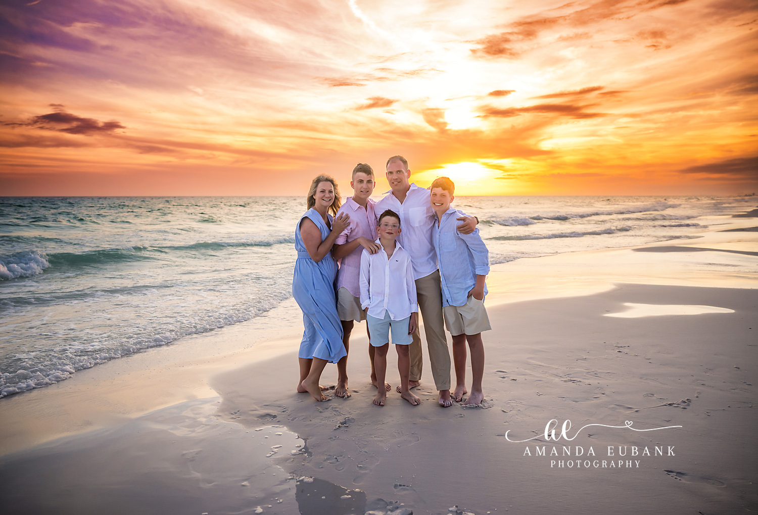 Seaside FL Family Hug Sunset Portrait | Gulf Surf Glow Family of five squeezed close together in coordinated pink, blue, and white linen as a glowing amber and lavender sunset blankets the Seaside, FL Gulf surf behind them.