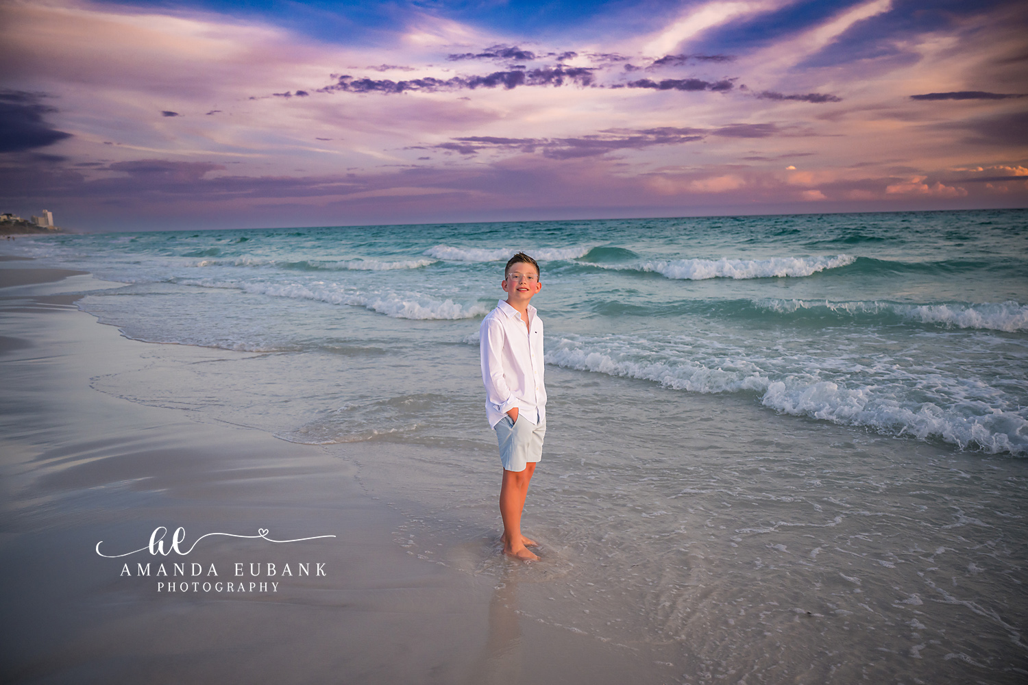 Seaside FL Little Boy Beach Portrait | Lavender Sunset Surf Smiling young boy in a white linen button-up and light blue shorts stands barefoot at the Seaside, FL surf line as a soft lavender and pink sunset glows above the rolling Gulf waves.