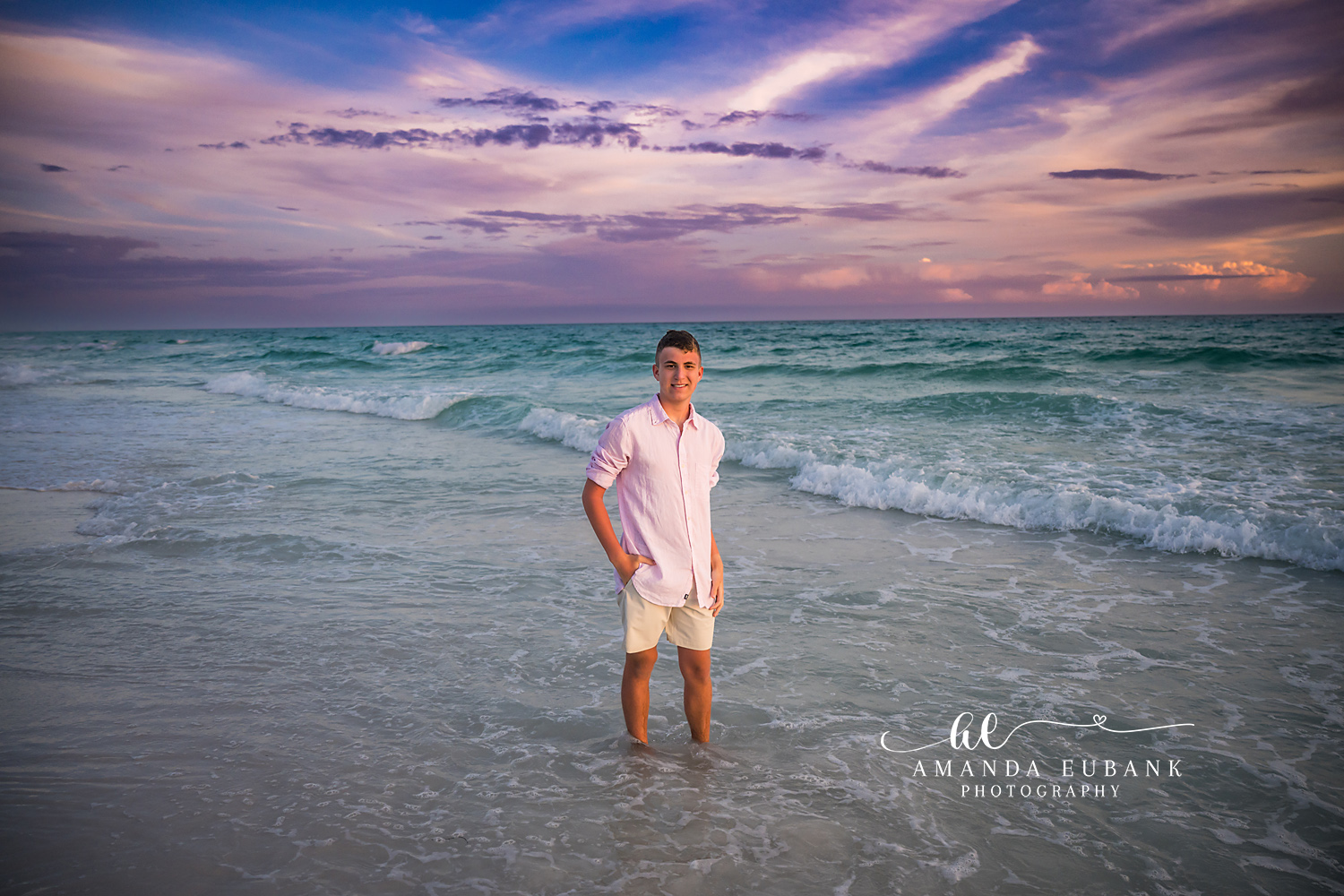 Seaside FL Senior Portrait | Young Man Standing in Gulf Surf Smiling teen senior in a soft pink button-up and cream shorts stands ankle-deep in the aqua Seaside, FL Gulf surf as soft lavender sunset clouds drift overhead.